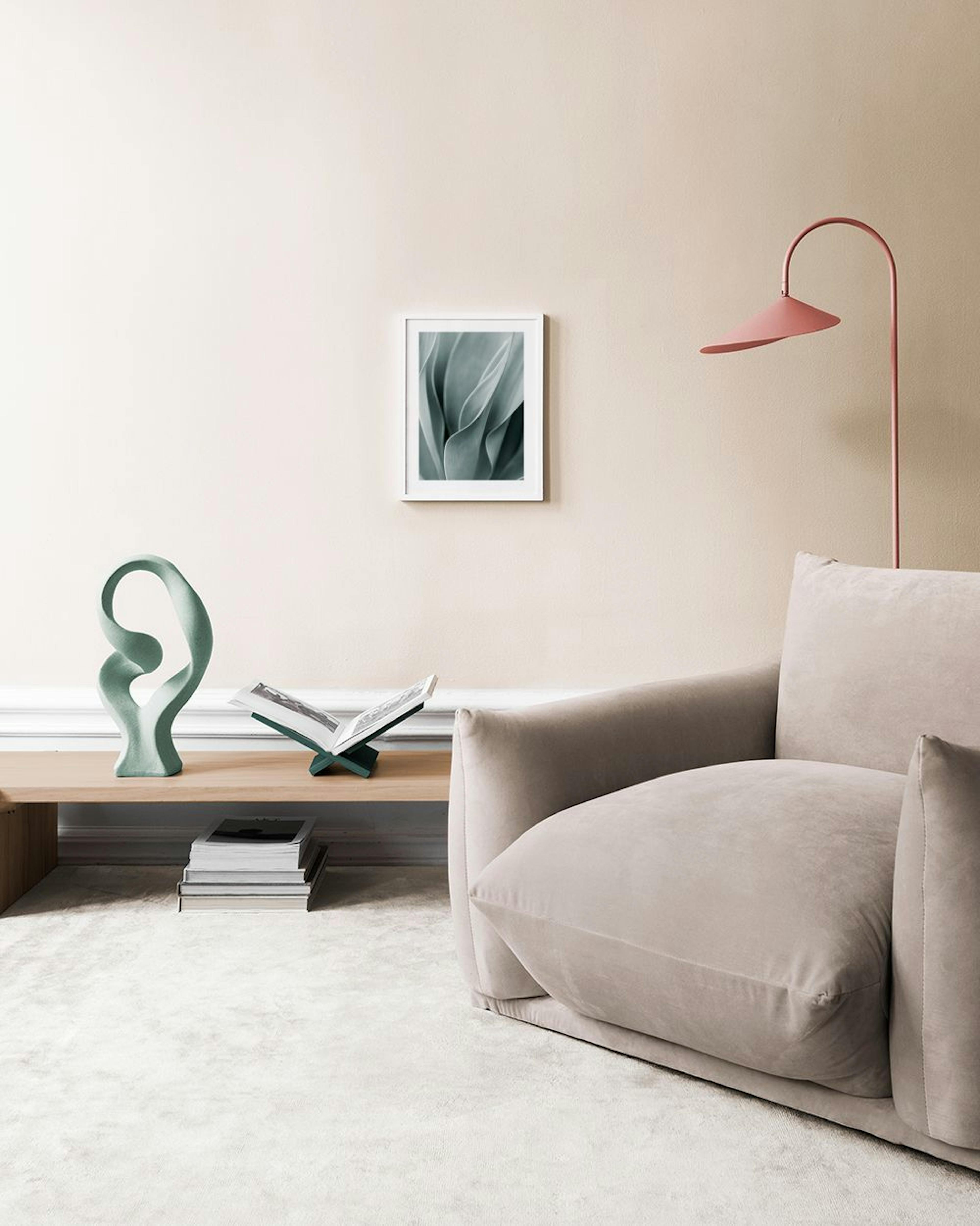 A print of soft green plant leaves with flowing lines, hanging above a natural wood and woven bench with books.