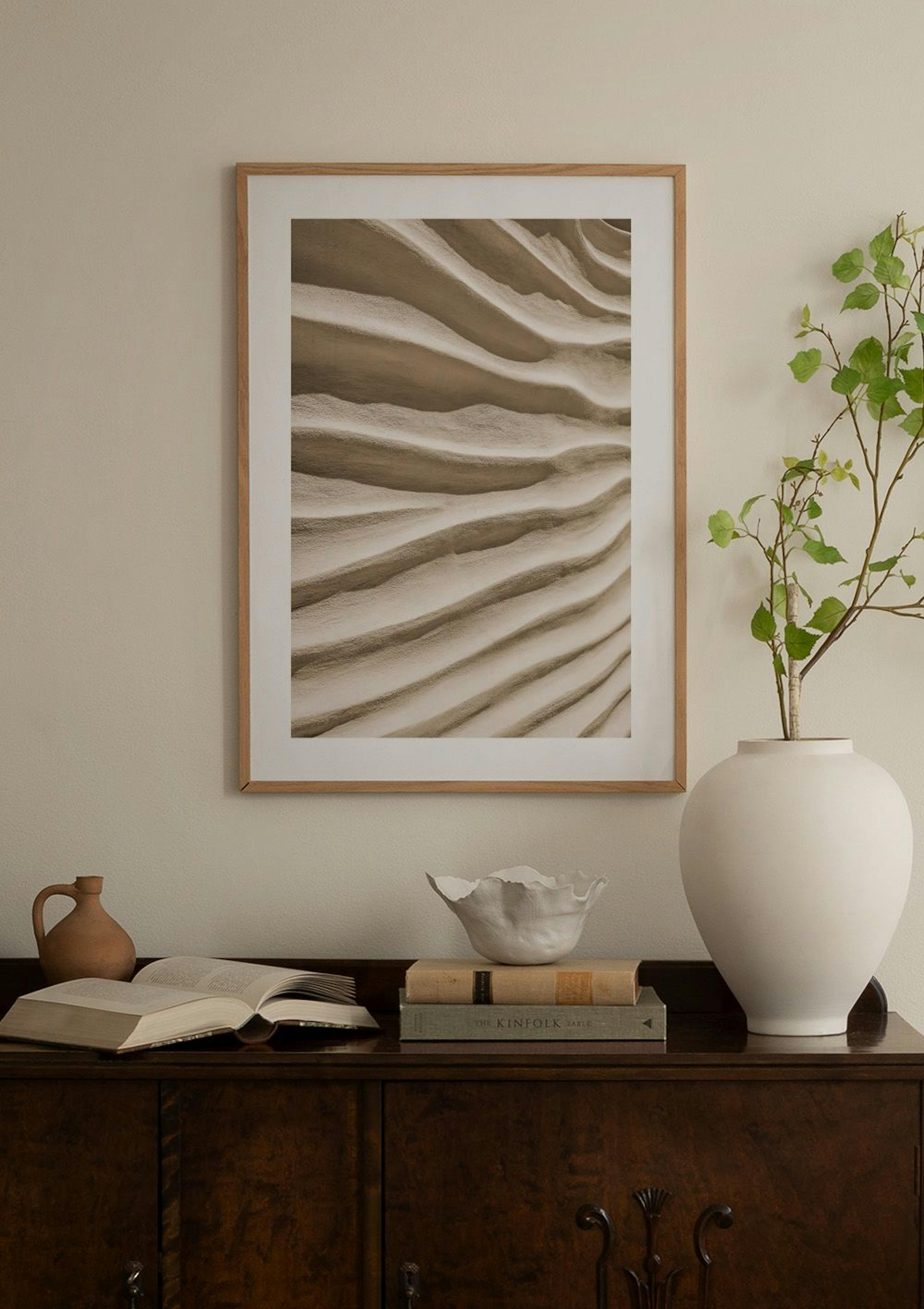 Poster mit beige-grauen Sanddünen in Wellenform, hängt über einer Ablage mit Buch und Vase.