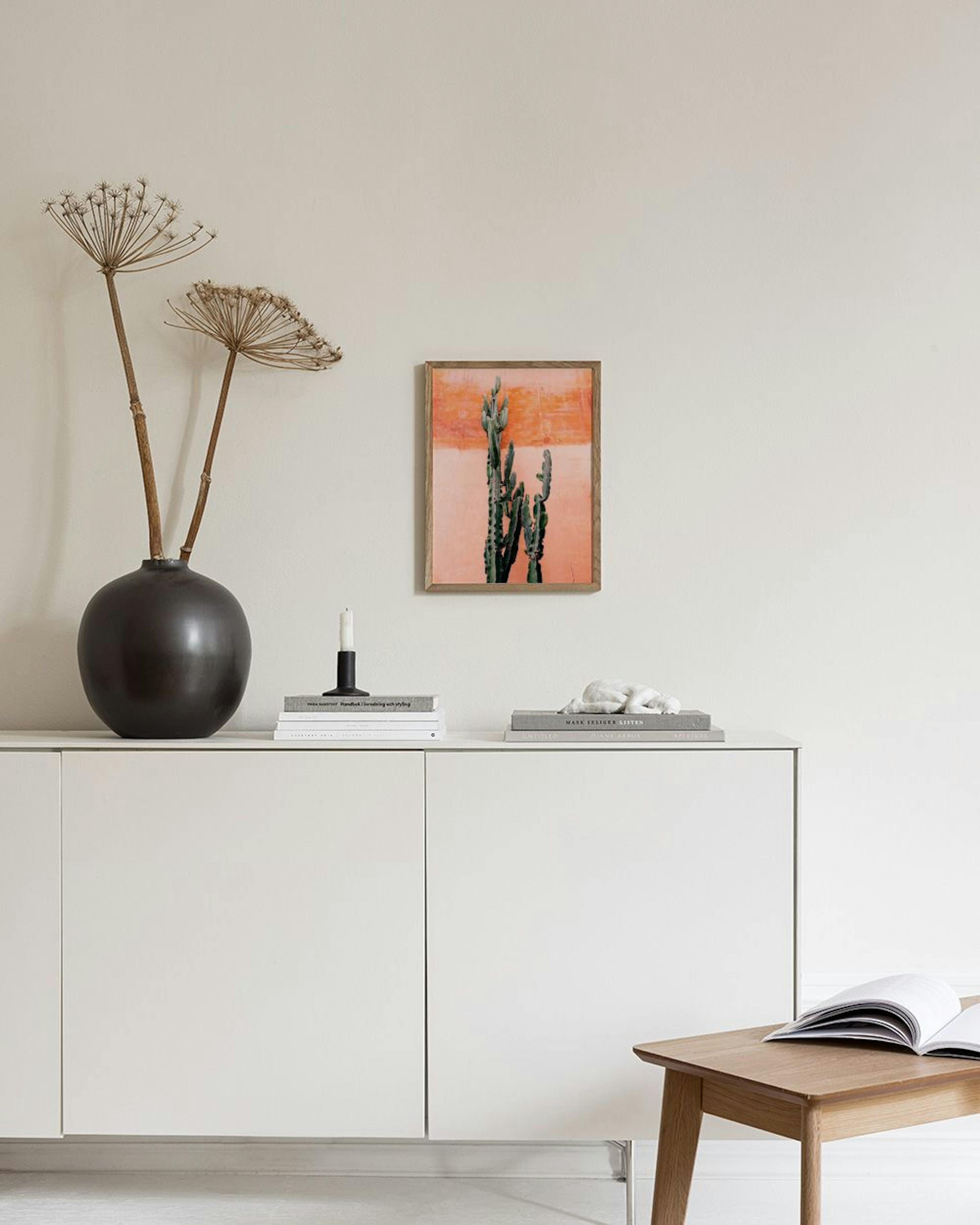 Poster mit grünem Kaktus vor einer strukturierten, orange-rosa Wand, über einem Beistelltisch mit Deko.