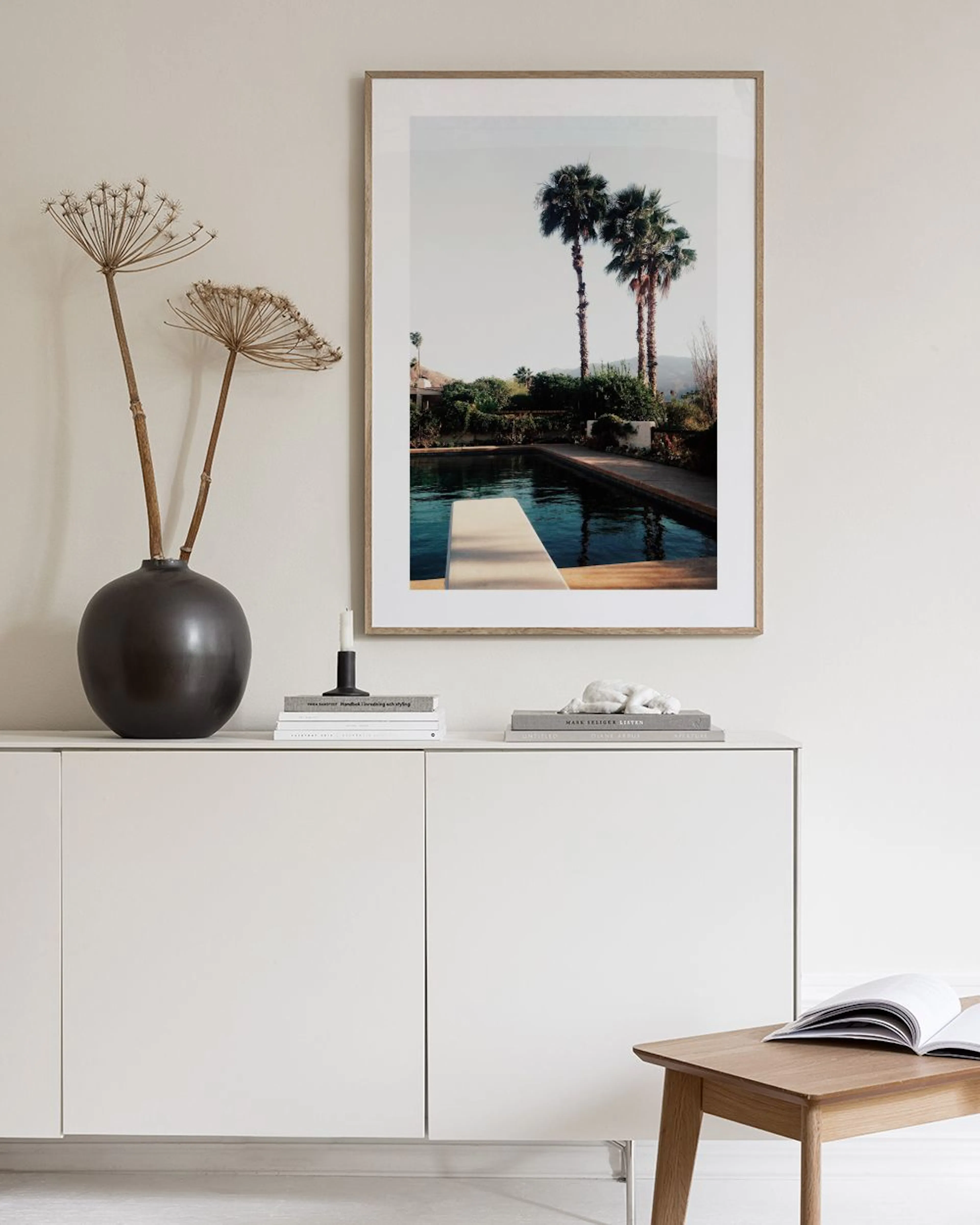 A print of a pool with palm trees and a diving board hanging above a wicker bench with books and glass vases.