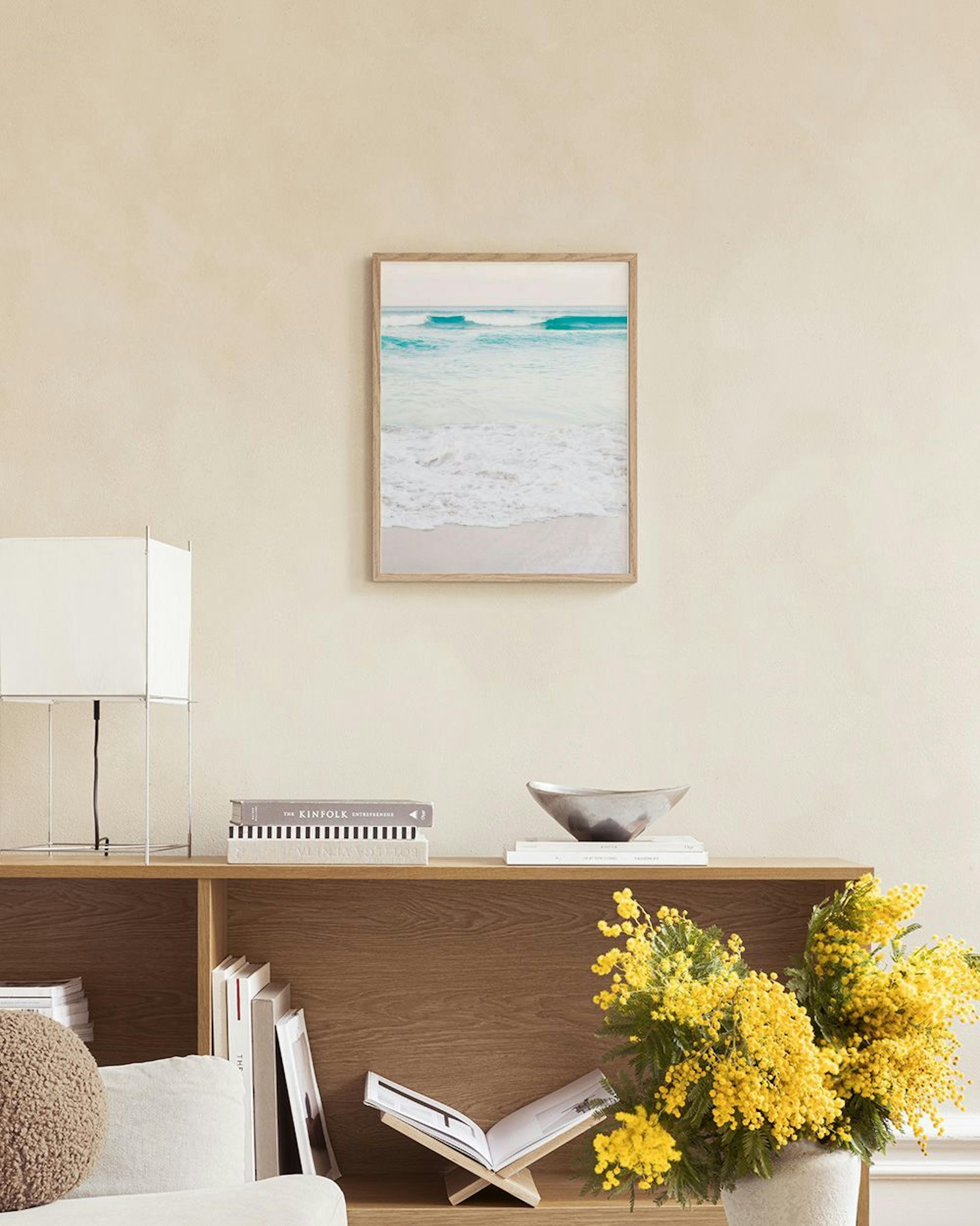 Póster de playa con olas turquesas y arena clara, colgado sobre un mueble blanco con un jarrón y libros.