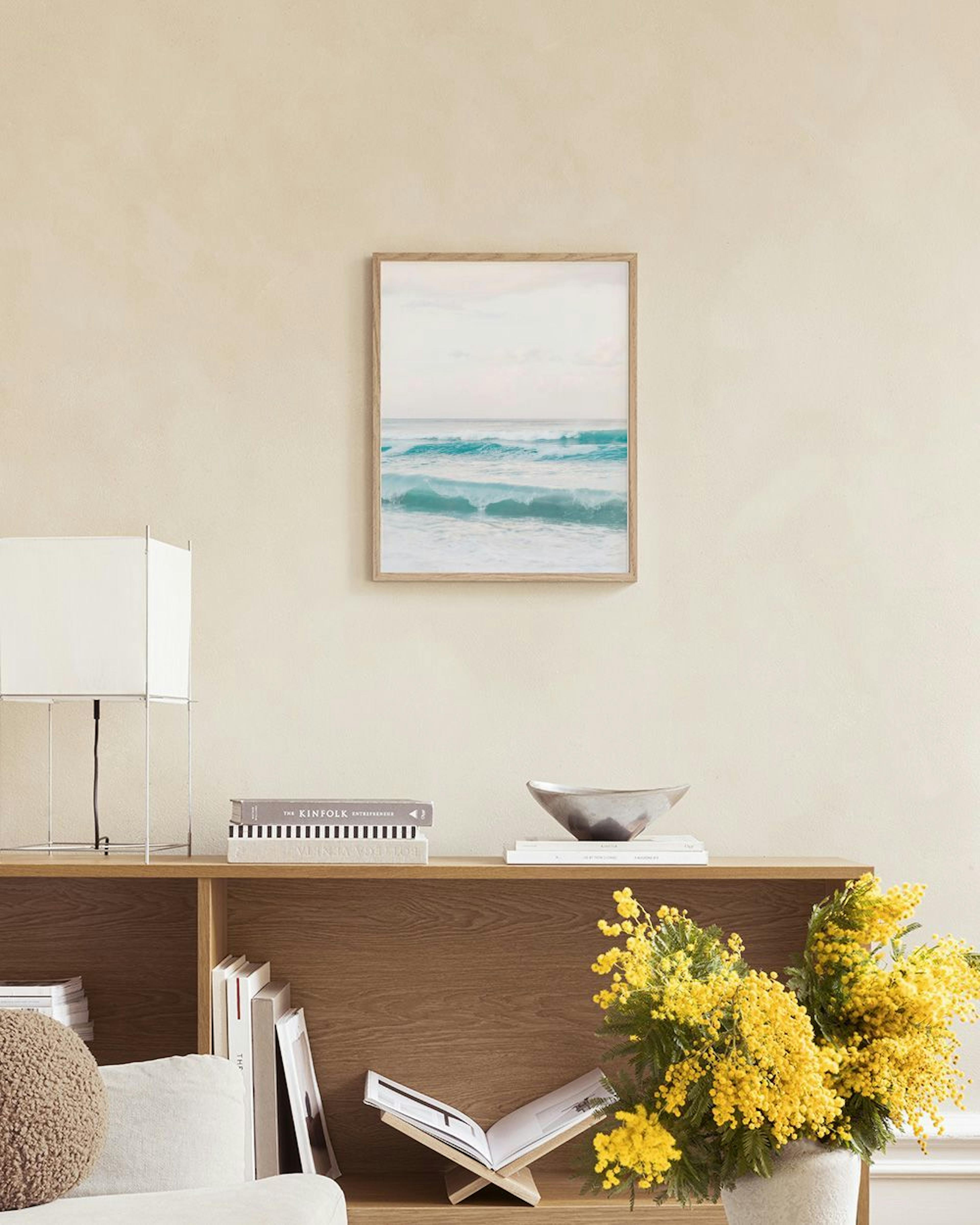 Un póster de olas turquesas del océano bajo un cielo gris, colgado sobre un mueble blanco con un libro abierto y un jarrón de cr