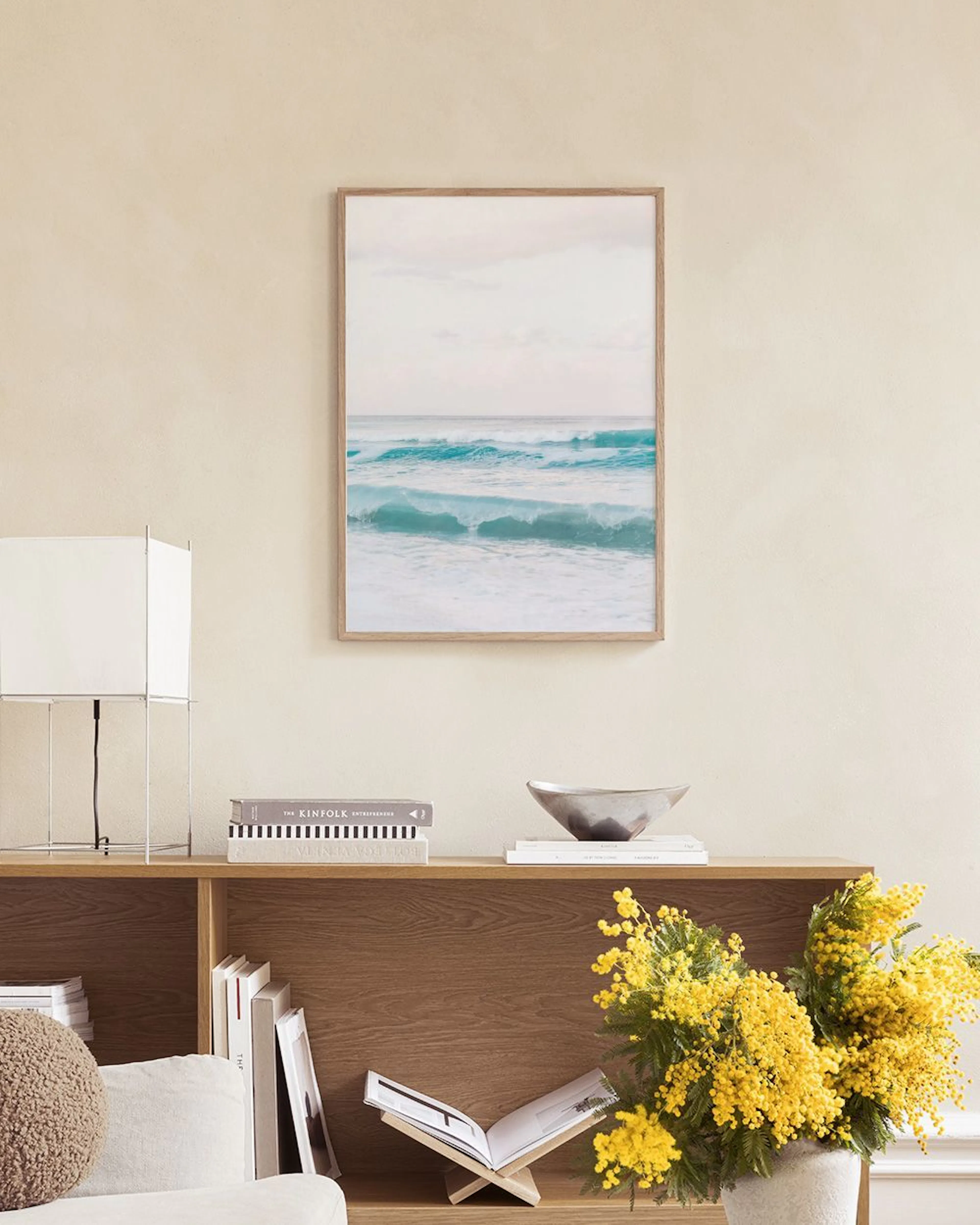 Un póster de olas turquesas del océano bajo un cielo gris, colgado sobre un mueble blanco con un libro abierto y un jarrón de cr