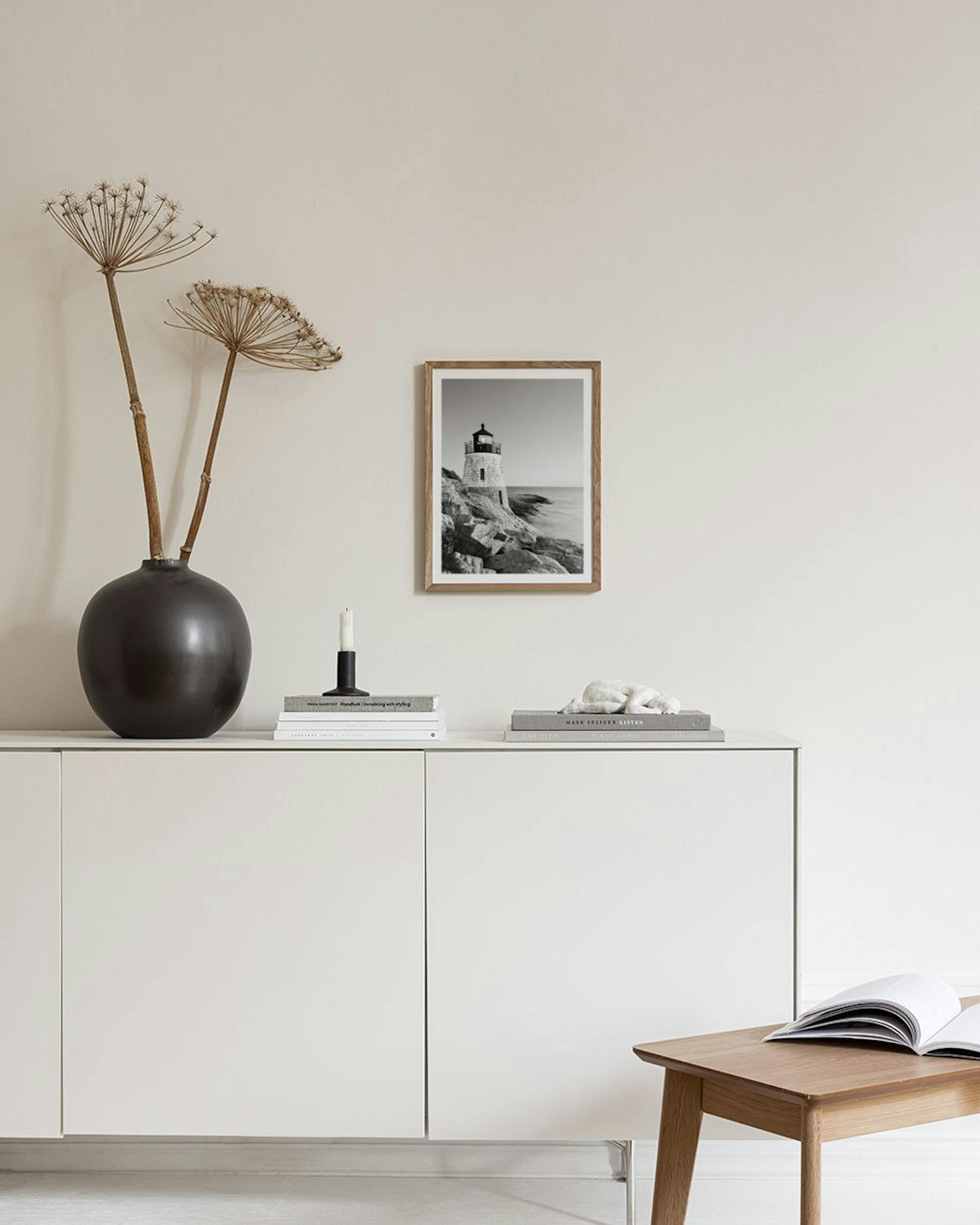 Póster de un faro de piedra en blanco y negro, sobre rocas junto al mar, exhibido en una repisa con objetos decorativos.