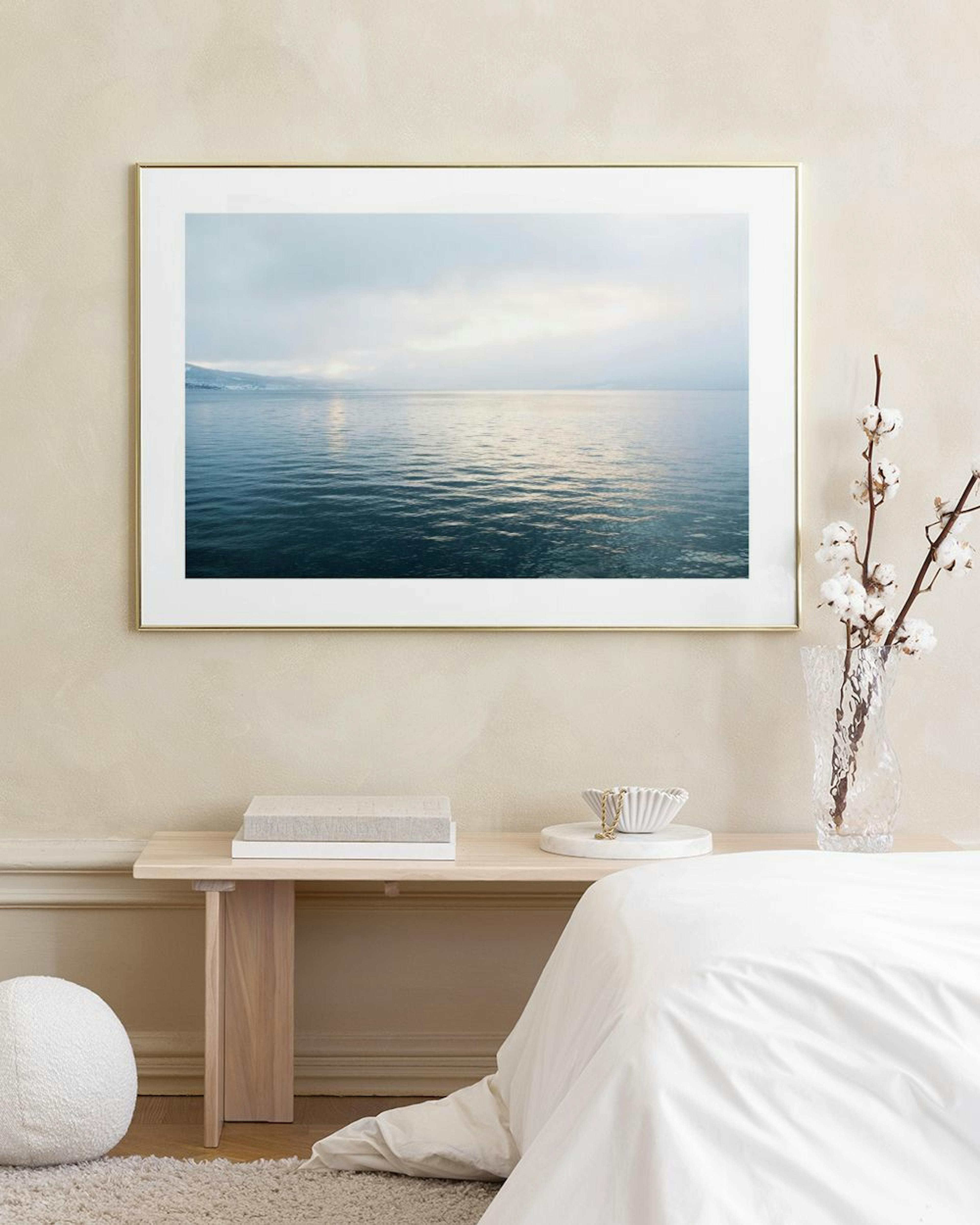 A print of a tranquil lake or ocean under a cloudy sky, hanging above a light wooden table with glasses.