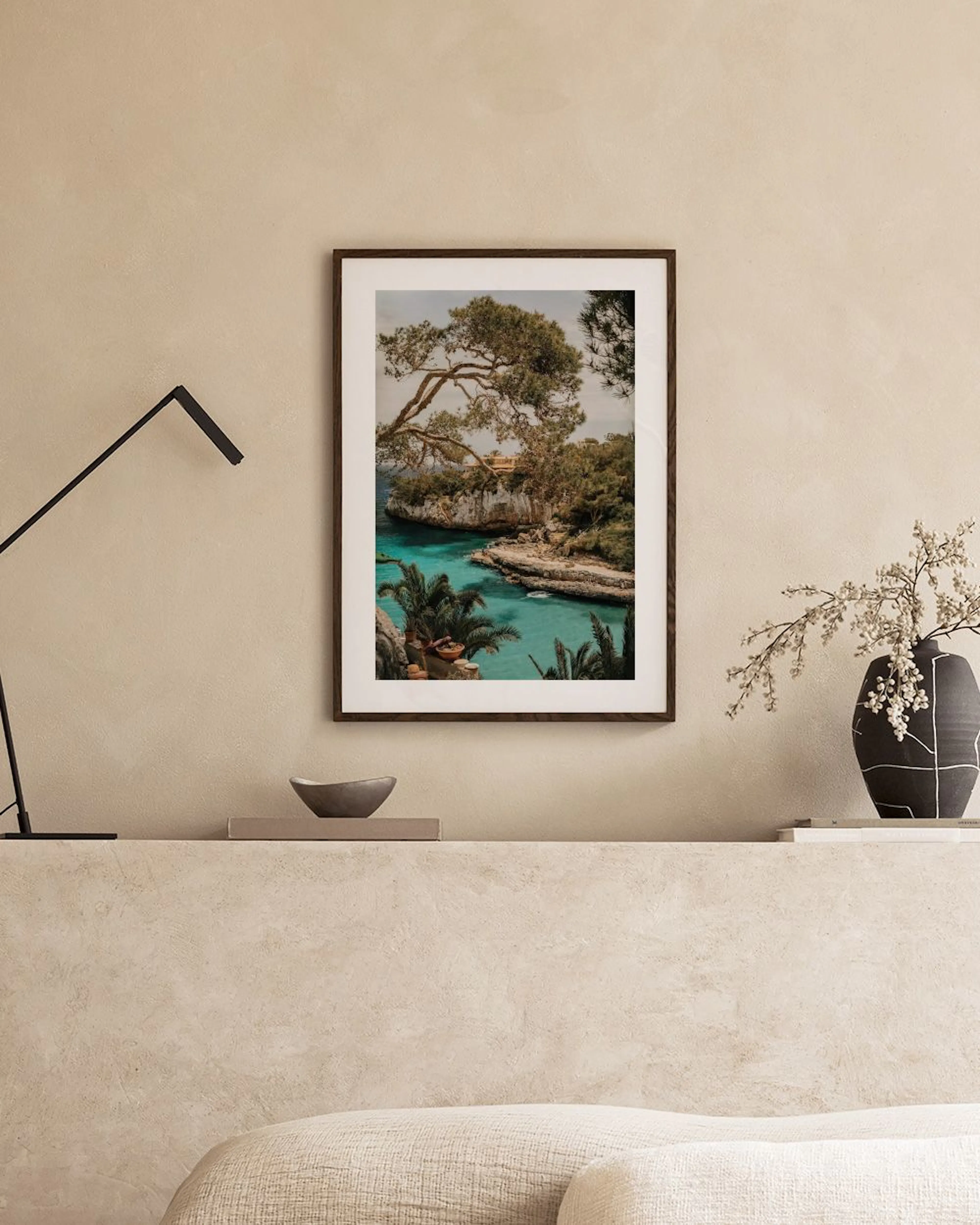 Póster de una cala de agua turquesa rodeada de árboles y acantilados, colgado sobre un sofá en una sala beige.