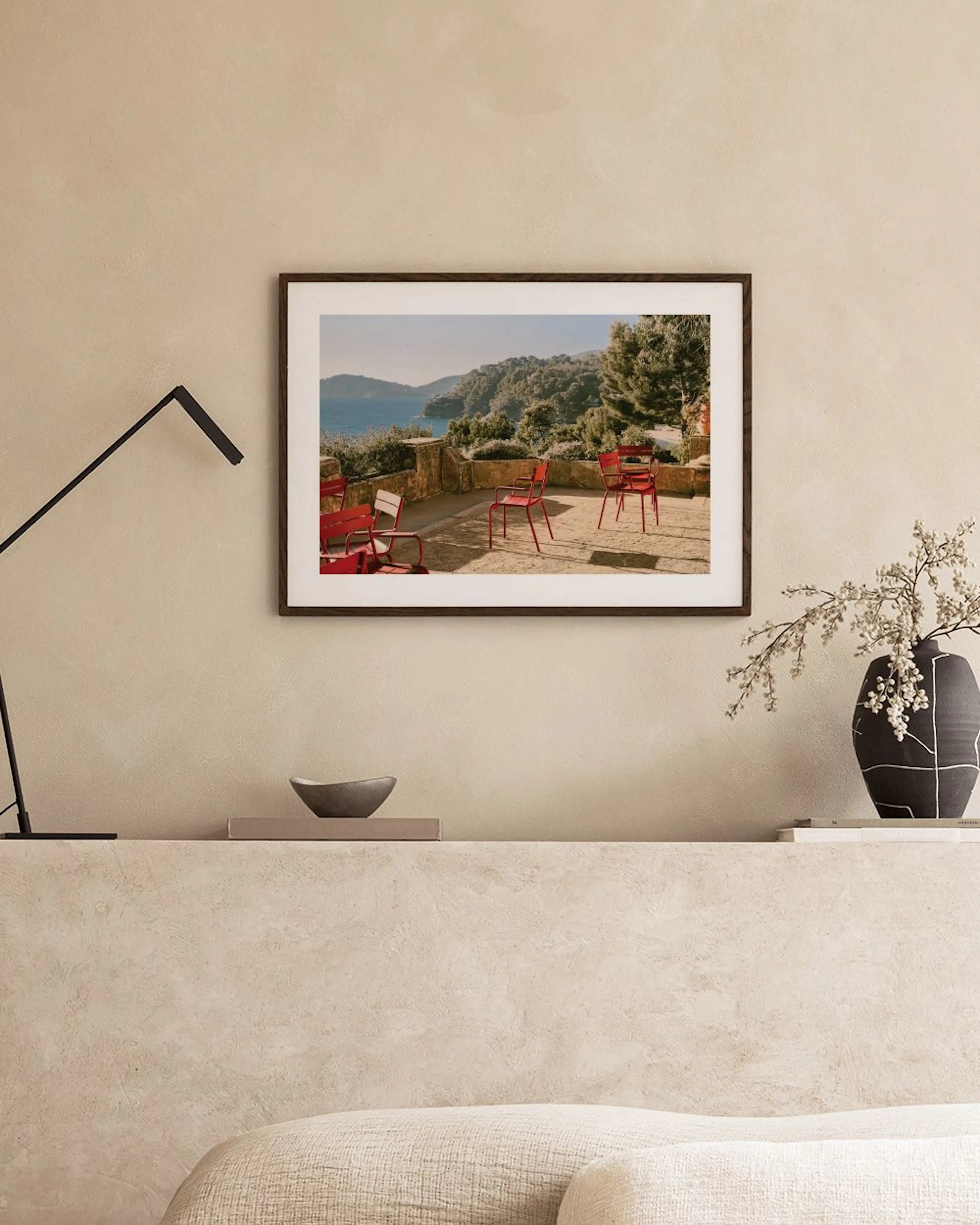 Poster eines mediterranen Ausblicks mit roten Stühlen auf einer Terrasse, hängend über einem beigefarbenen Sofa.