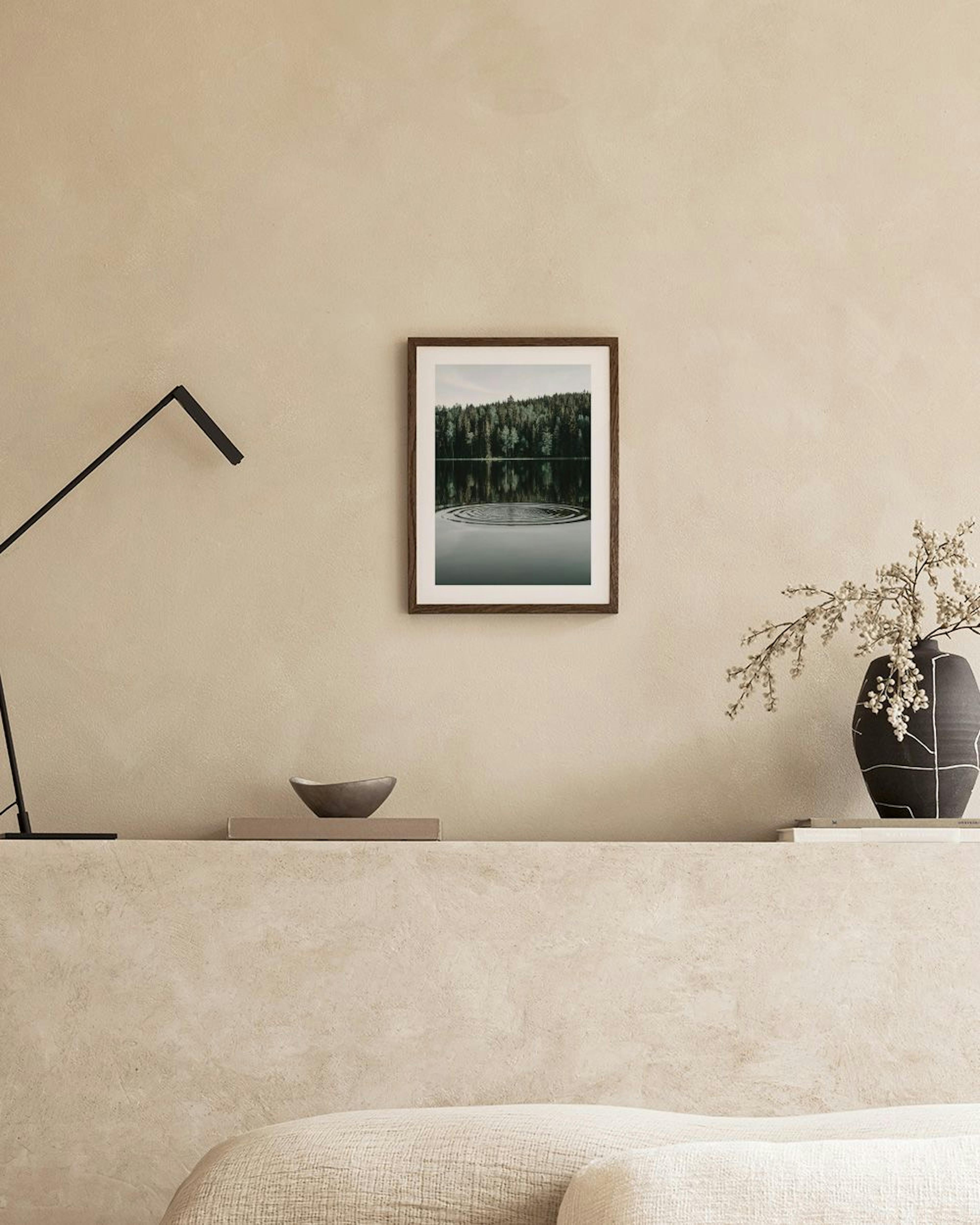 En plakat af en skov, der spejler sig i et stille vand med ringe på overfladen, hængende på en grå væg over et sidebord med vase