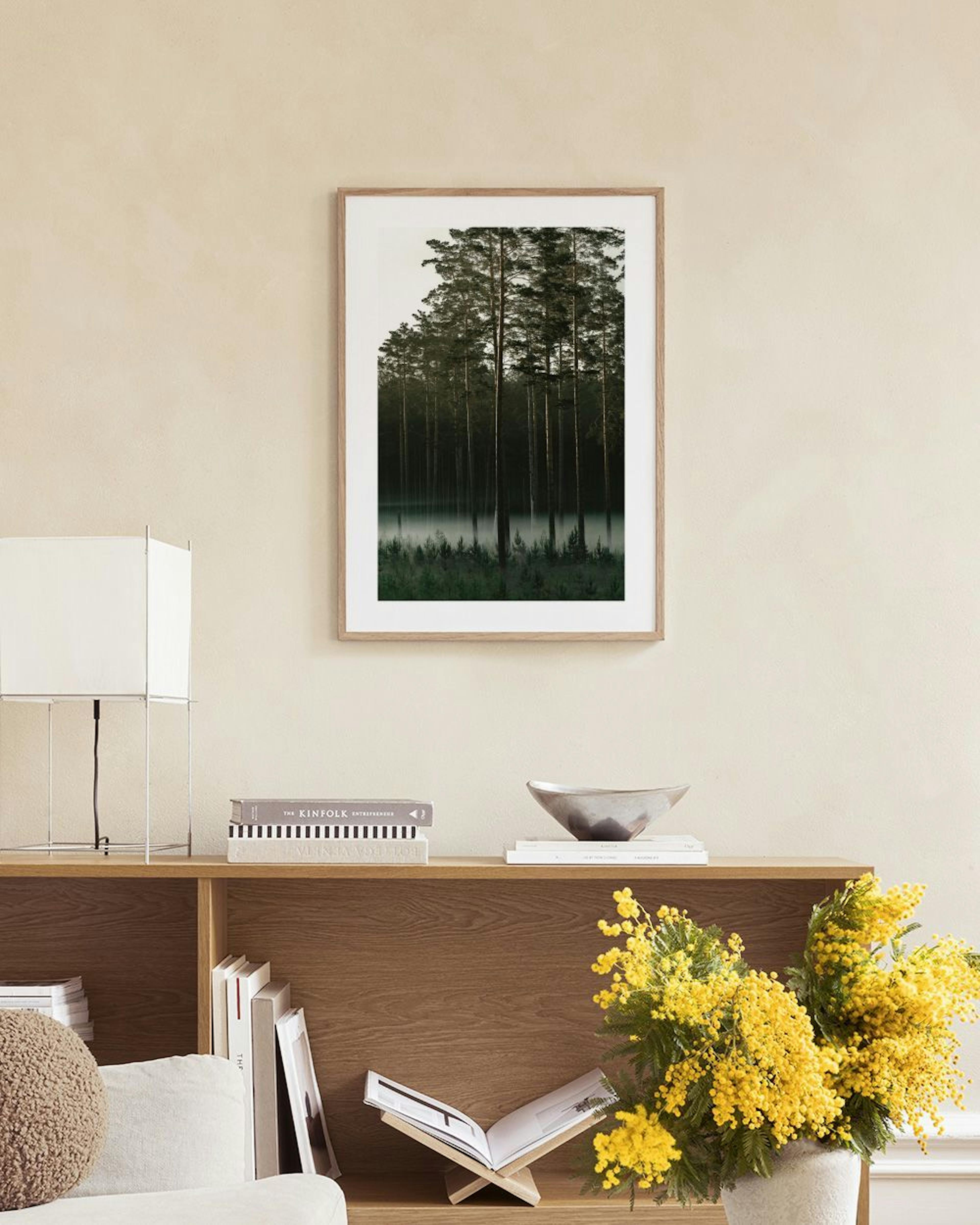 Póster de un bosque brumoso con pinos de color verde oscuro, colgando en una sala moderna sobre un libro abierto y jarrones.