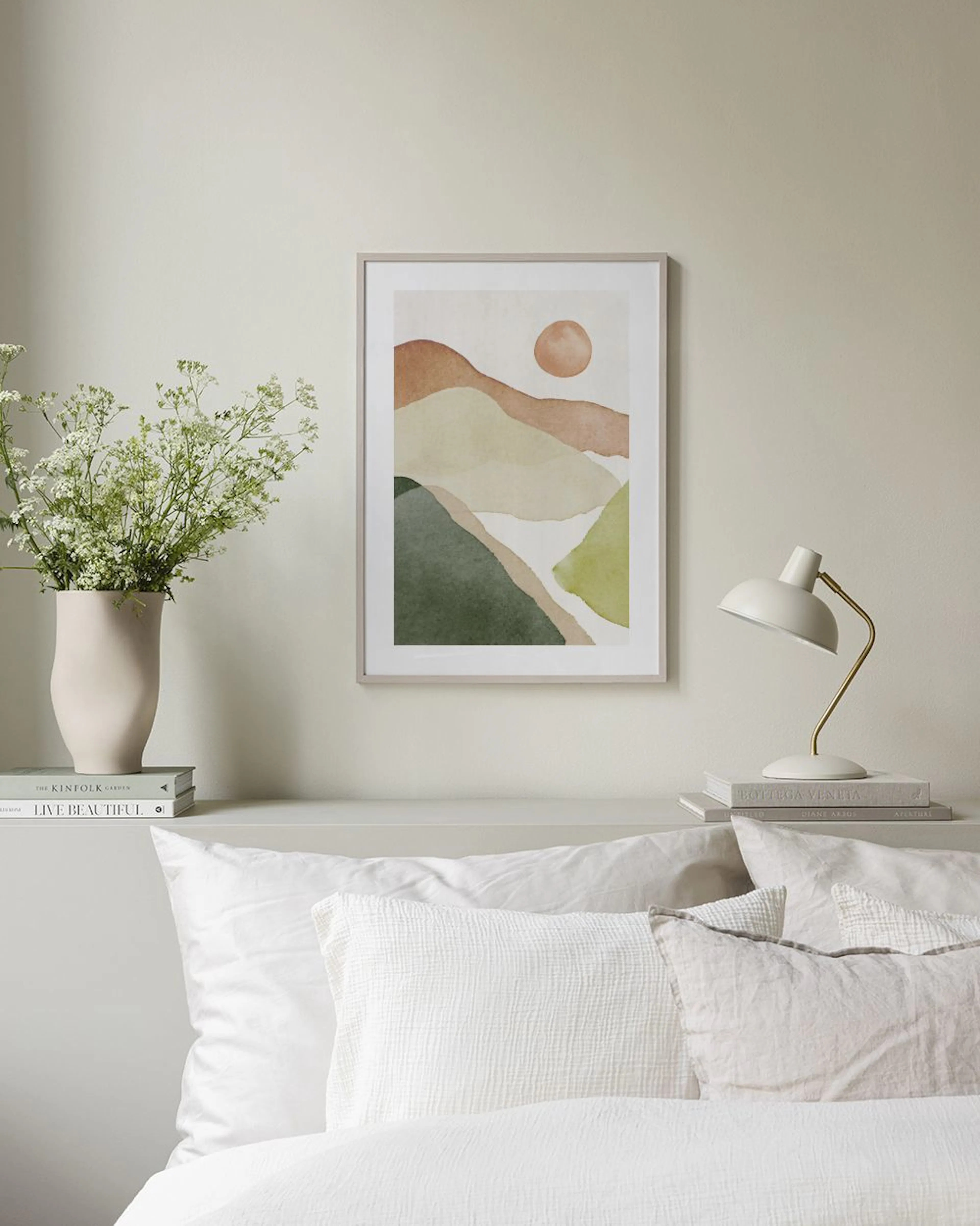 Plakat med akvarellfjell i grønt, brunt og beige, med en oker sol, henger over et hvitt skap med vase og bok.