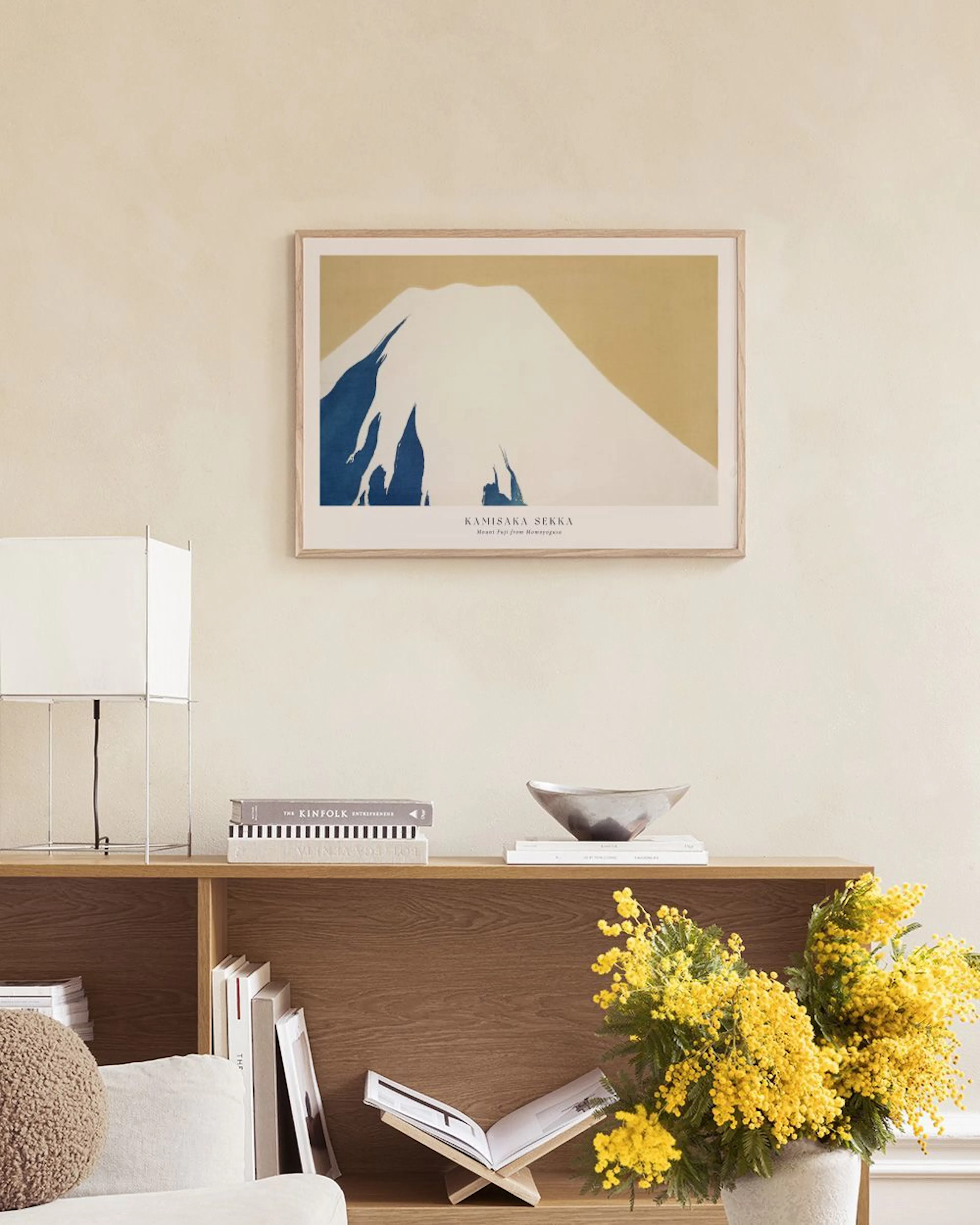 Póster de Kamisa Senka con el Monte Fuji sobre fondo salmón, colgado sobre un mueble blanco con libros y una planta.