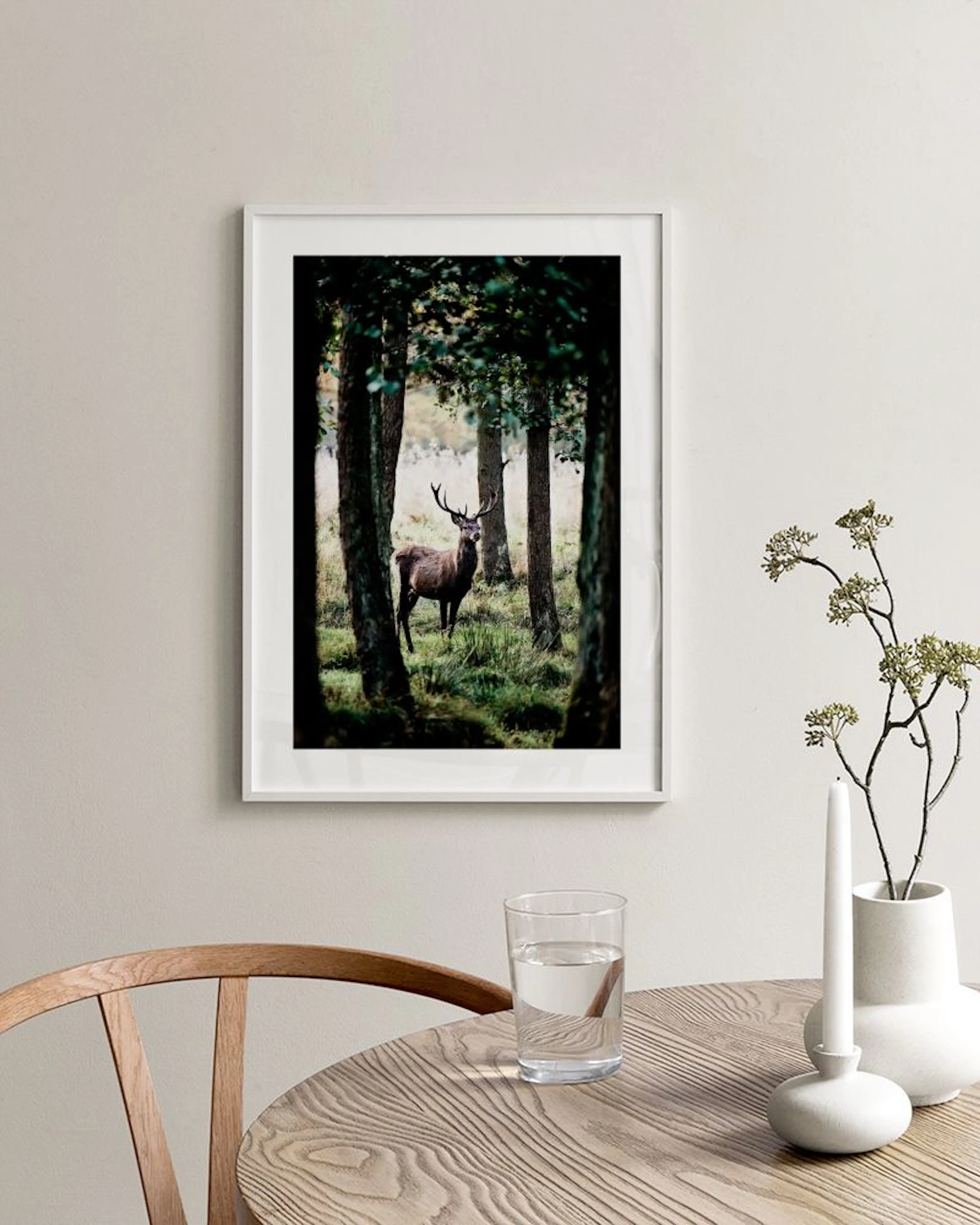 Poster van een hert in een mistig bos met donkere bomen en groen gras, hangend boven een wit dressoir in een woonkamer.