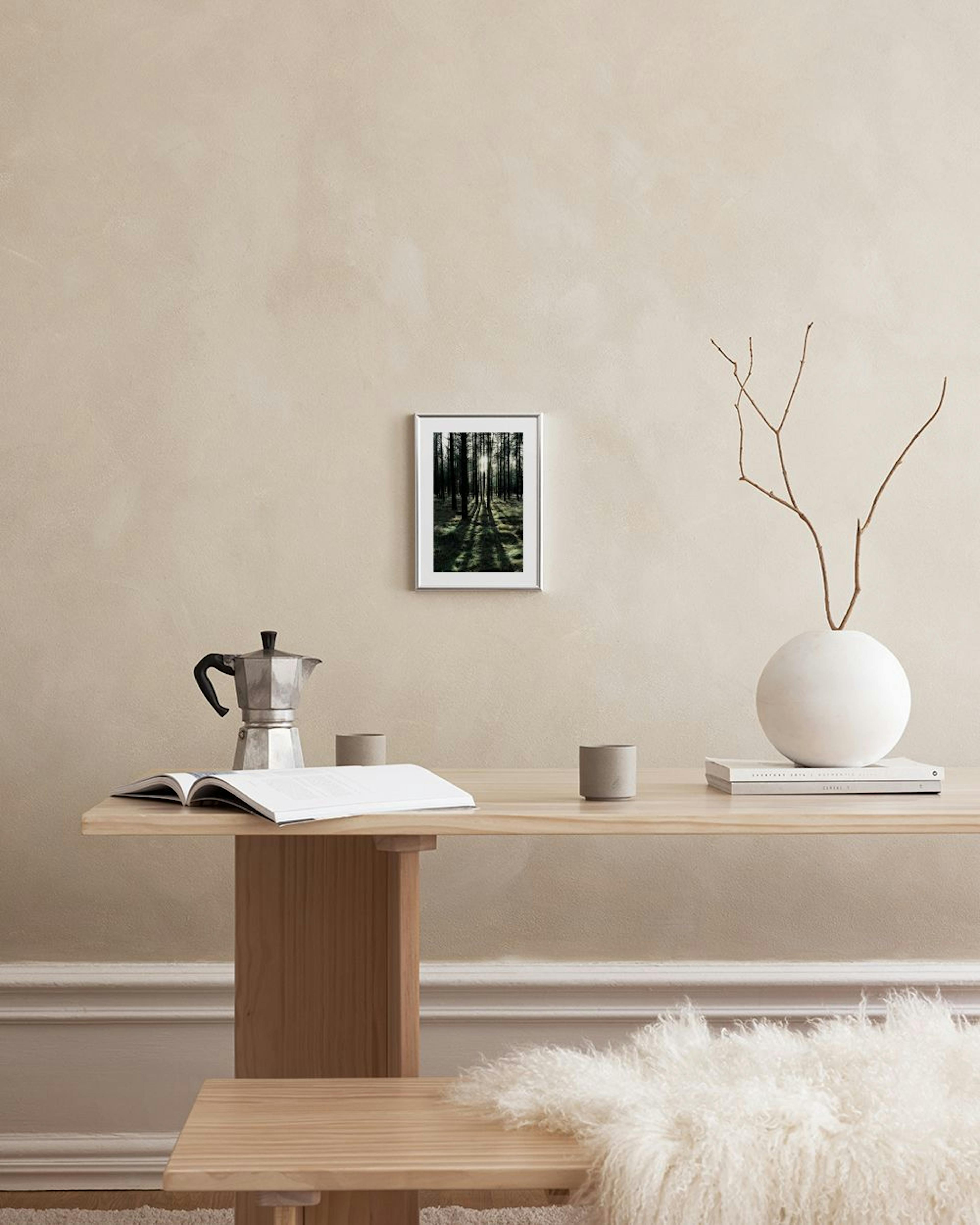 A print of a dark forest with sunbeams and long shadows, hanging on a white wall above a stool with books.