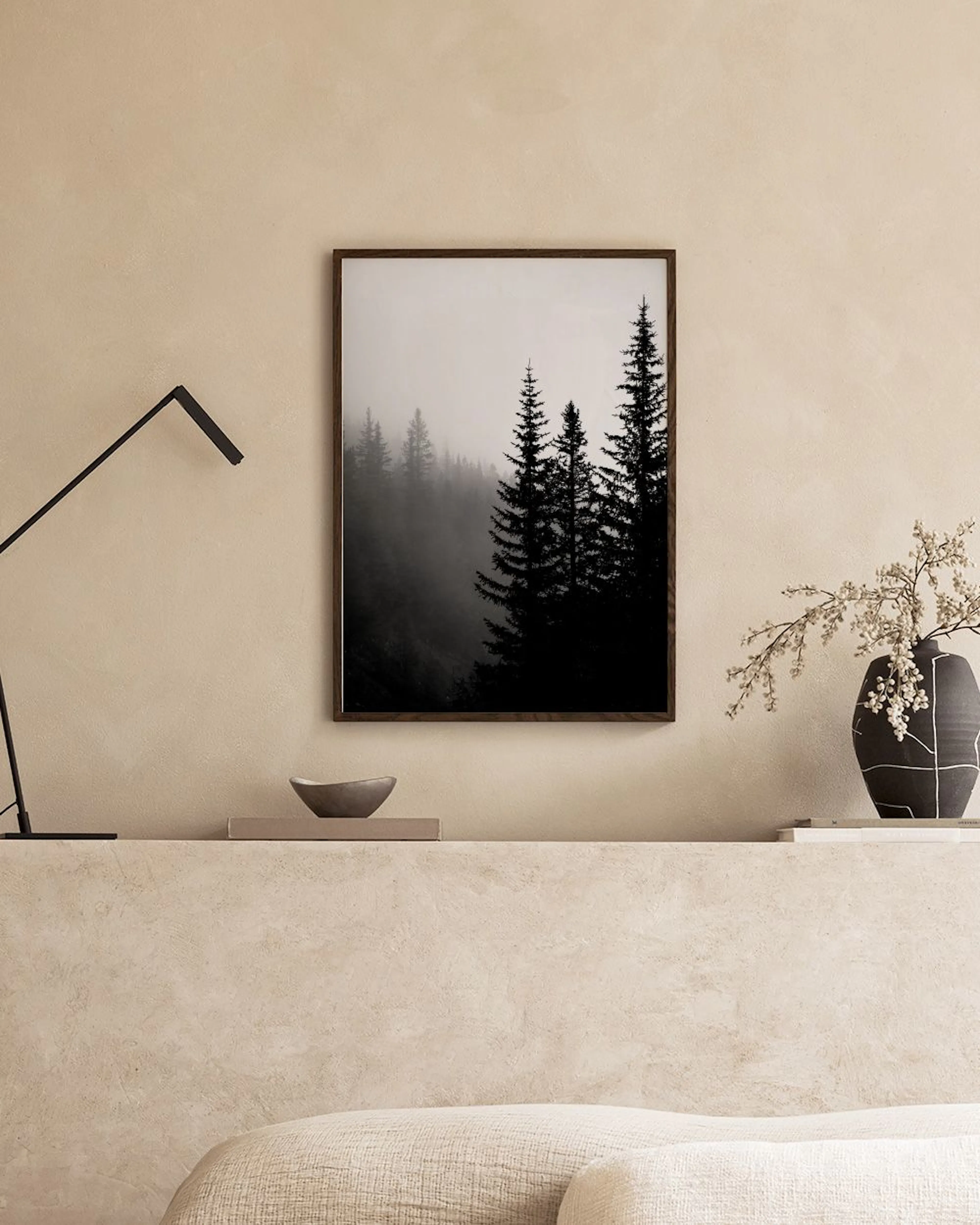 Poster van donkere naaldbomen in mistig bos, hangend boven een wit dressoir met boeken en planten.