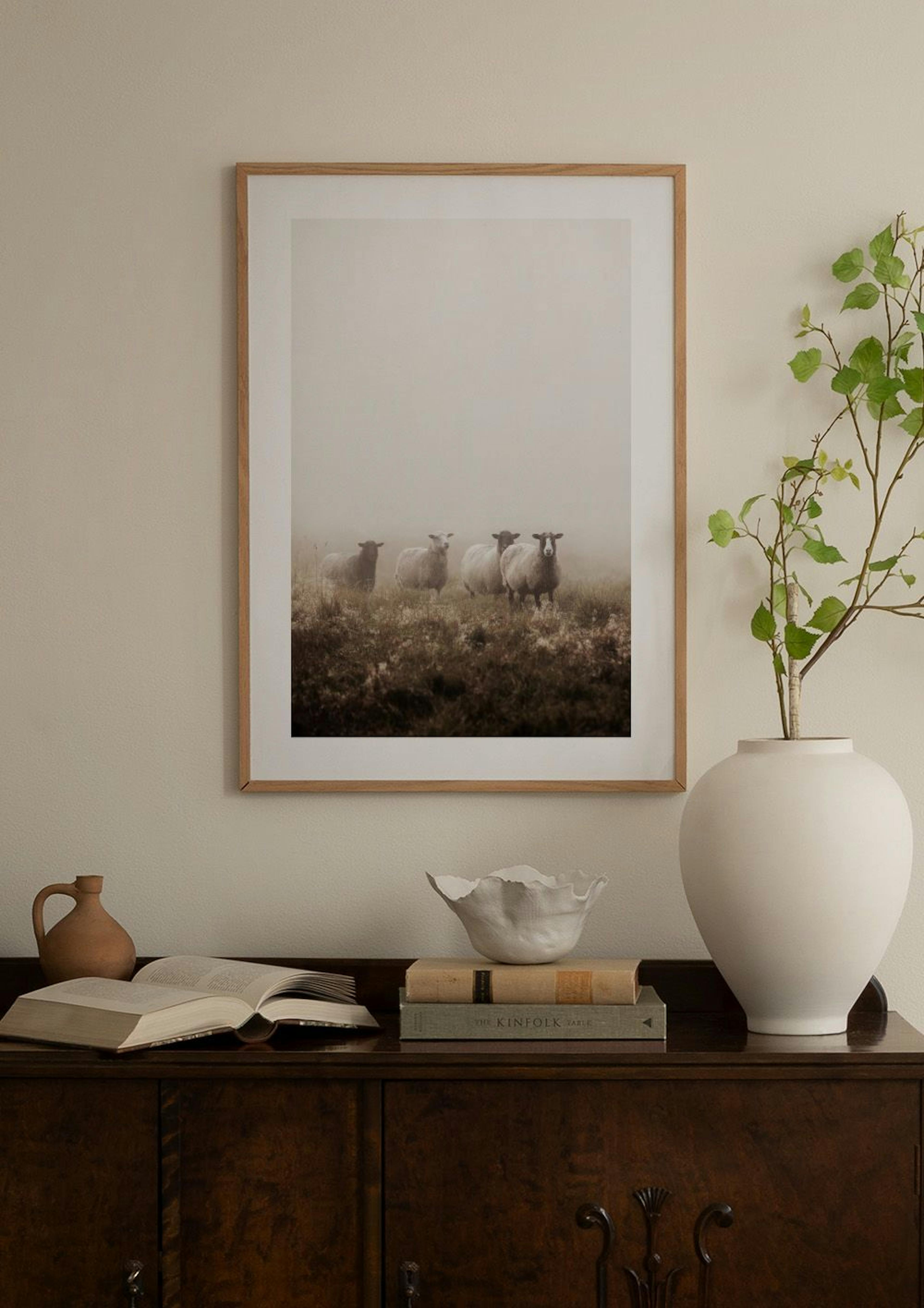 Póster de ovejas en un campo cubierto de niebla, colgado sobre una mesa auxiliar con una lámpara y un puf en un salón.