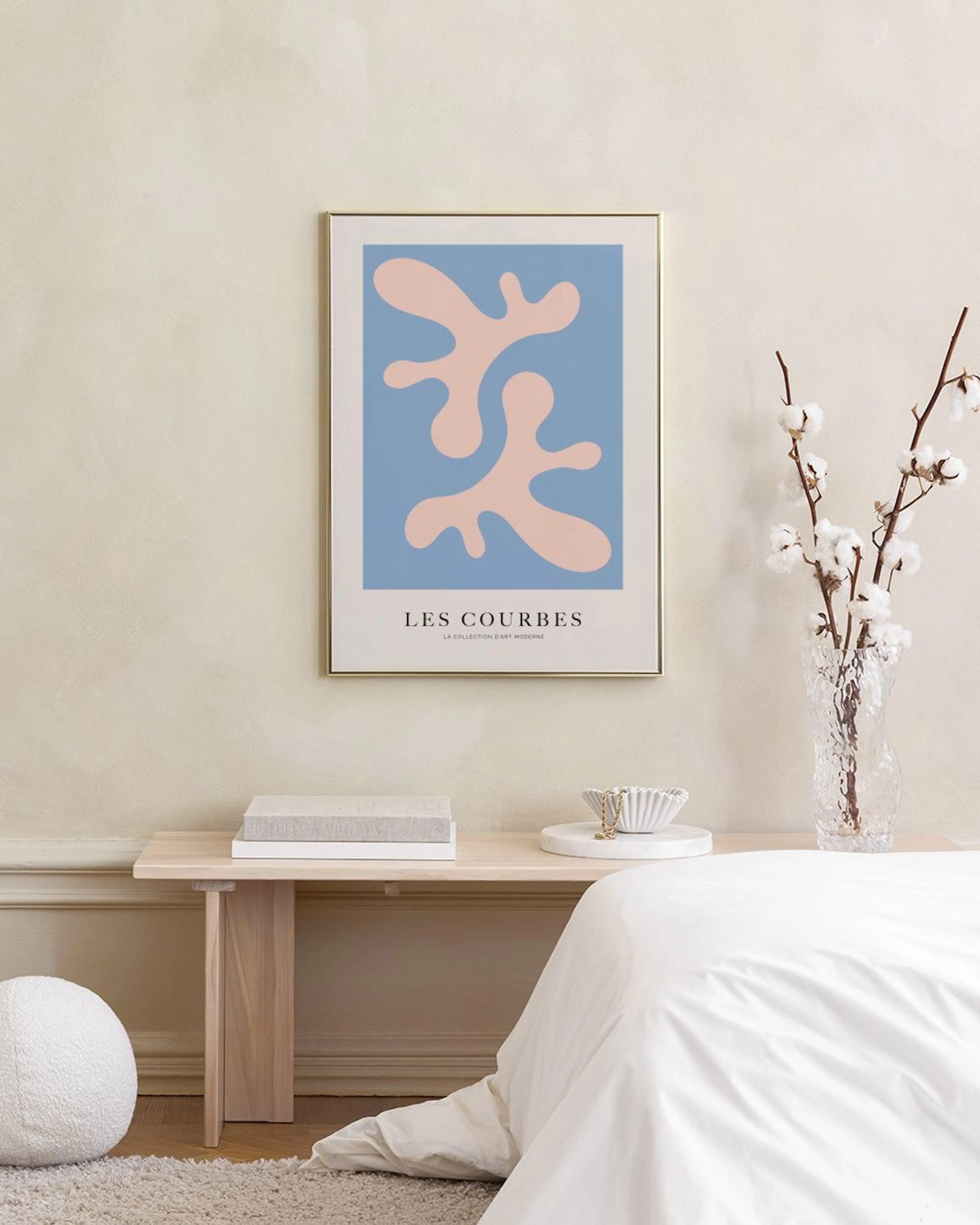 Poster med rosa organiska former mot blå bakgrund, hänger på en grå vägg ovanför ett mörkblått sammetstyg.