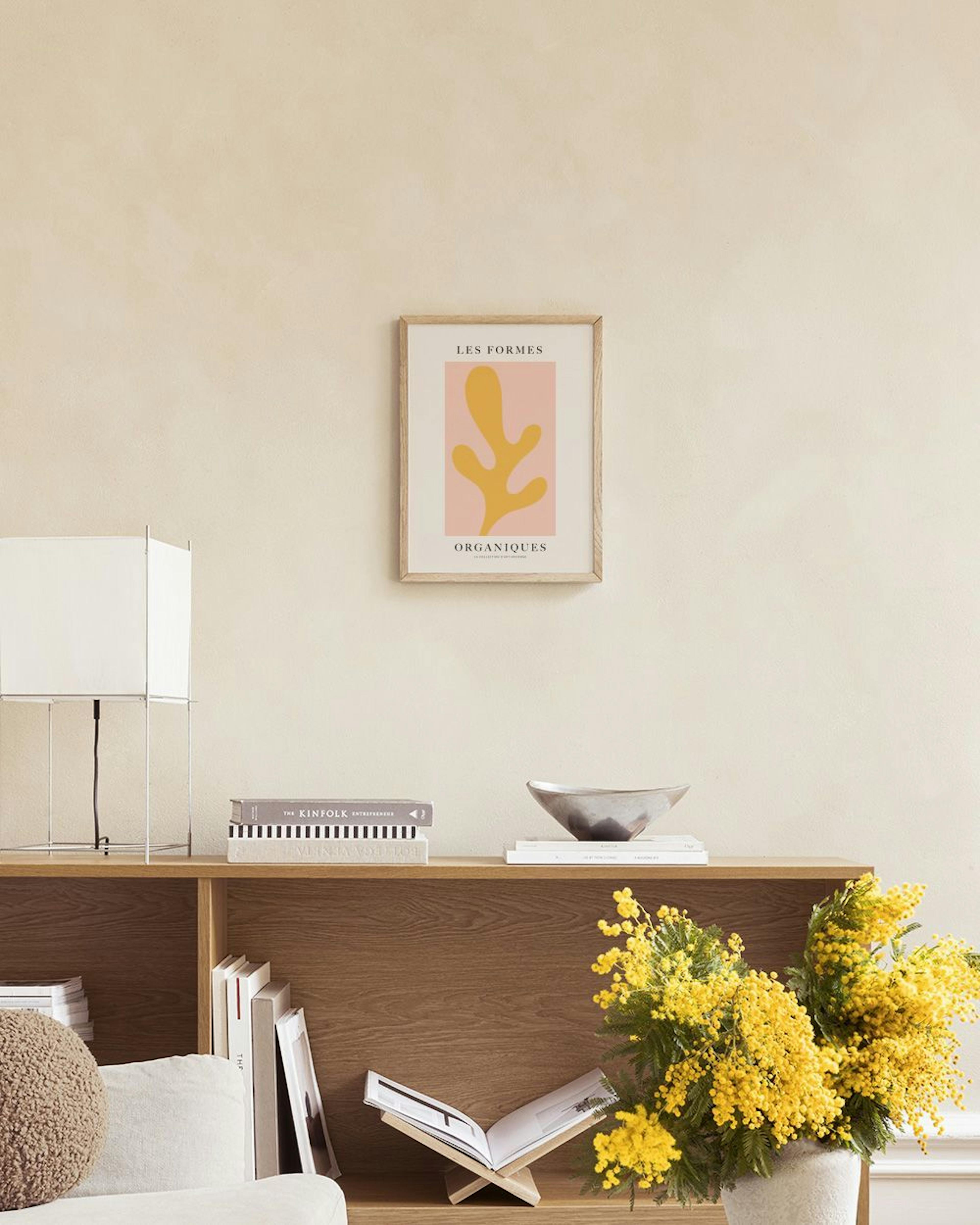 Póster de formas orgánicas amarillas sobre fondo rosa claro, colgado sobre un mueble blanco con libros y un jarrón con plumas.
