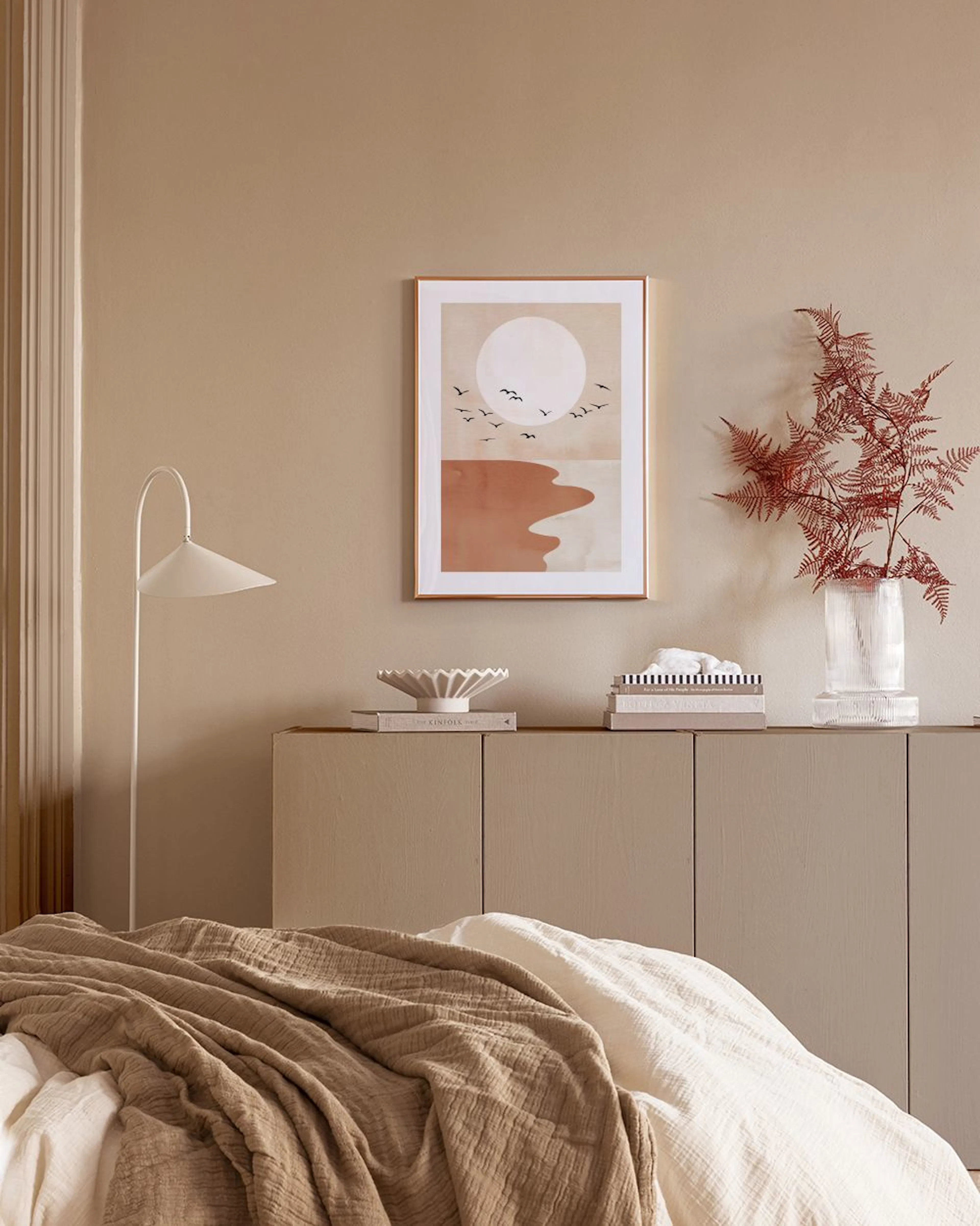A print of a minimalist desert landscape with a large white sun and flying birds, hanging in a modern room with pampas grass and