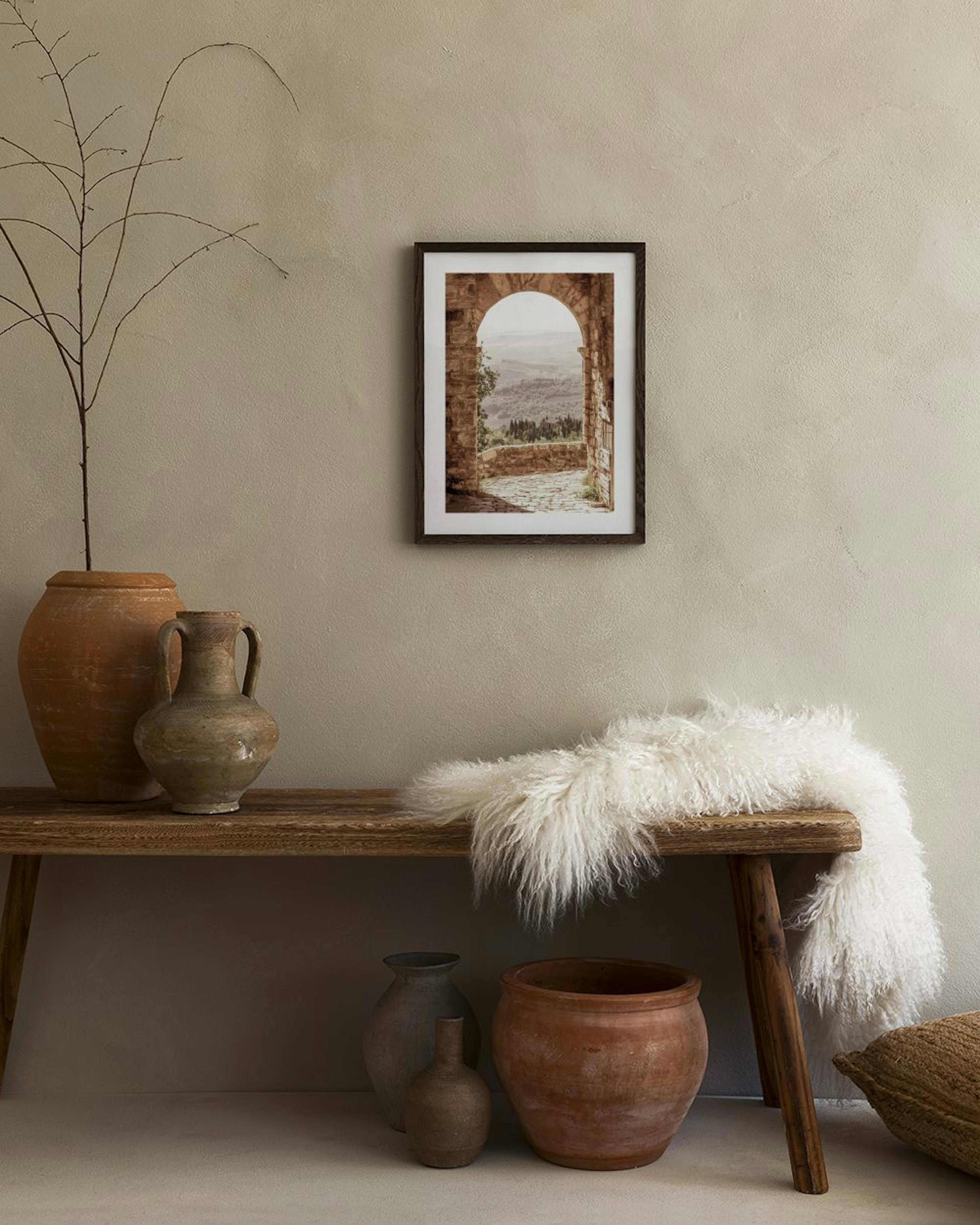 Un póster de un arco de piedra con vista a un paisaje montañoso, colgado sobre un mueble blanco con un florero y un libro.