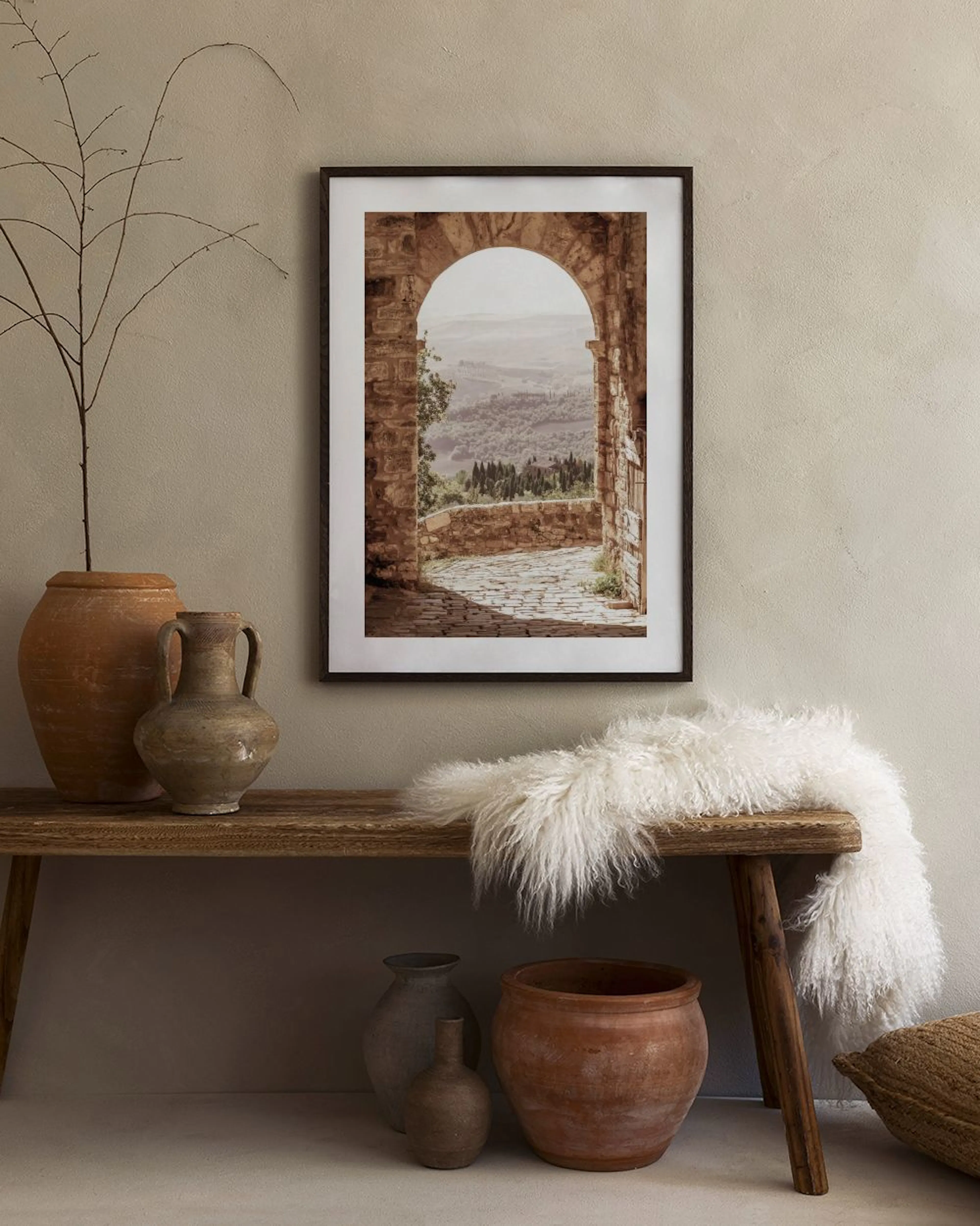 Poster van een stenen boog met uitzicht op een heuvelachtig landschap, boven een wit dressoir met vaas en open boek.