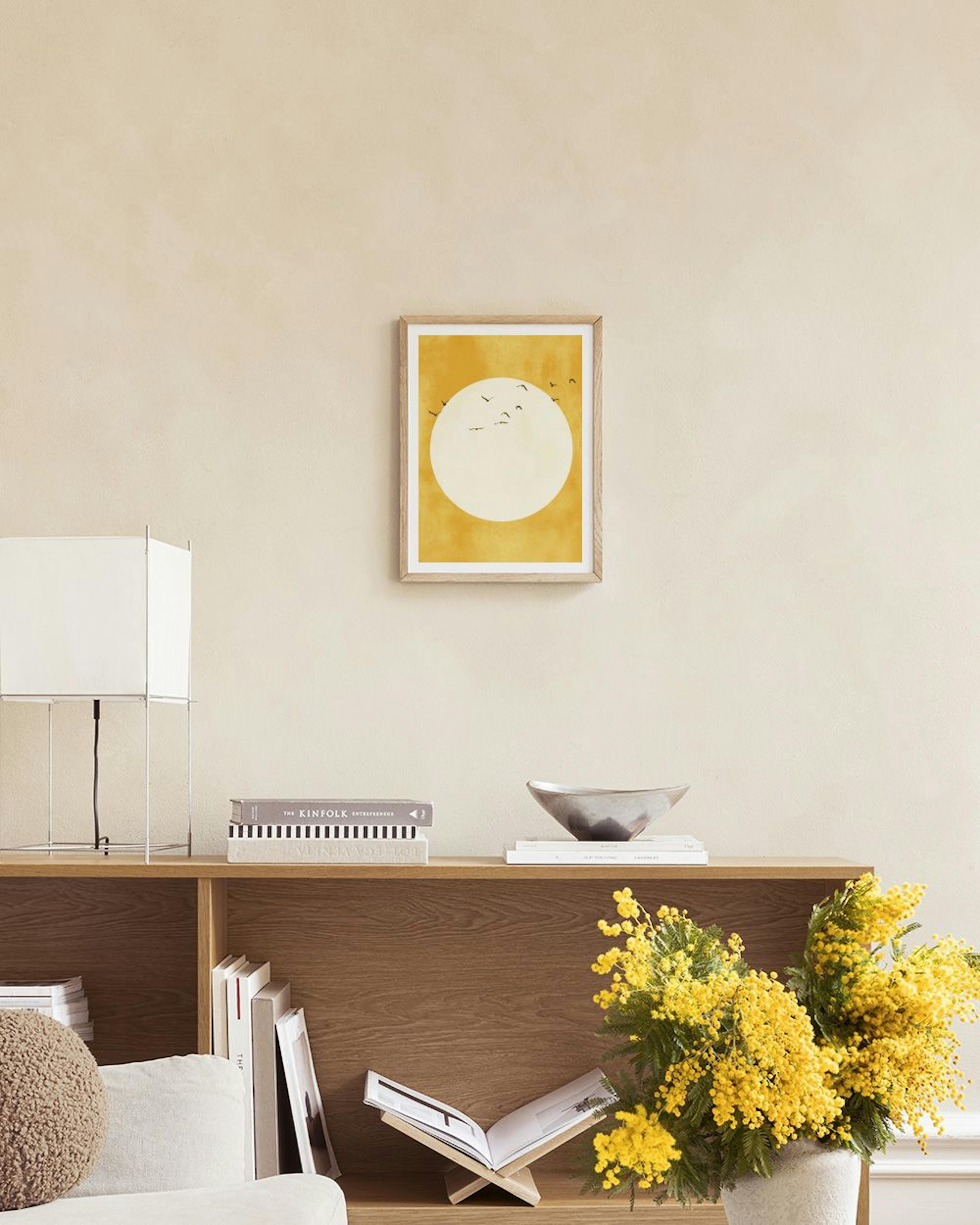 A print of a golden sun with flying birds, hanging above a gray velvet chair and a black side table in a room.