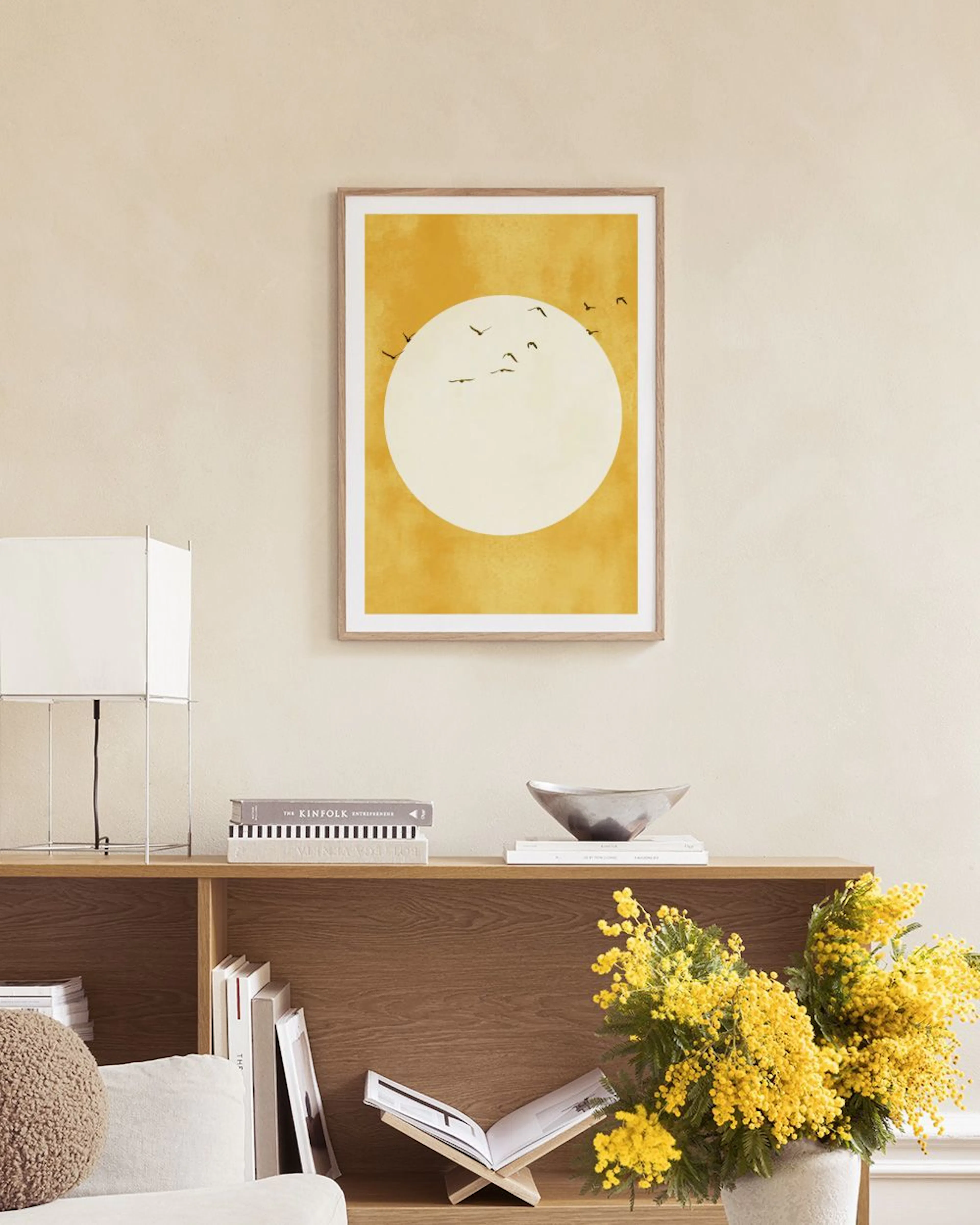 A print of a golden sun with flying birds, hanging above a gray chair and a black side table with a decorative vase.