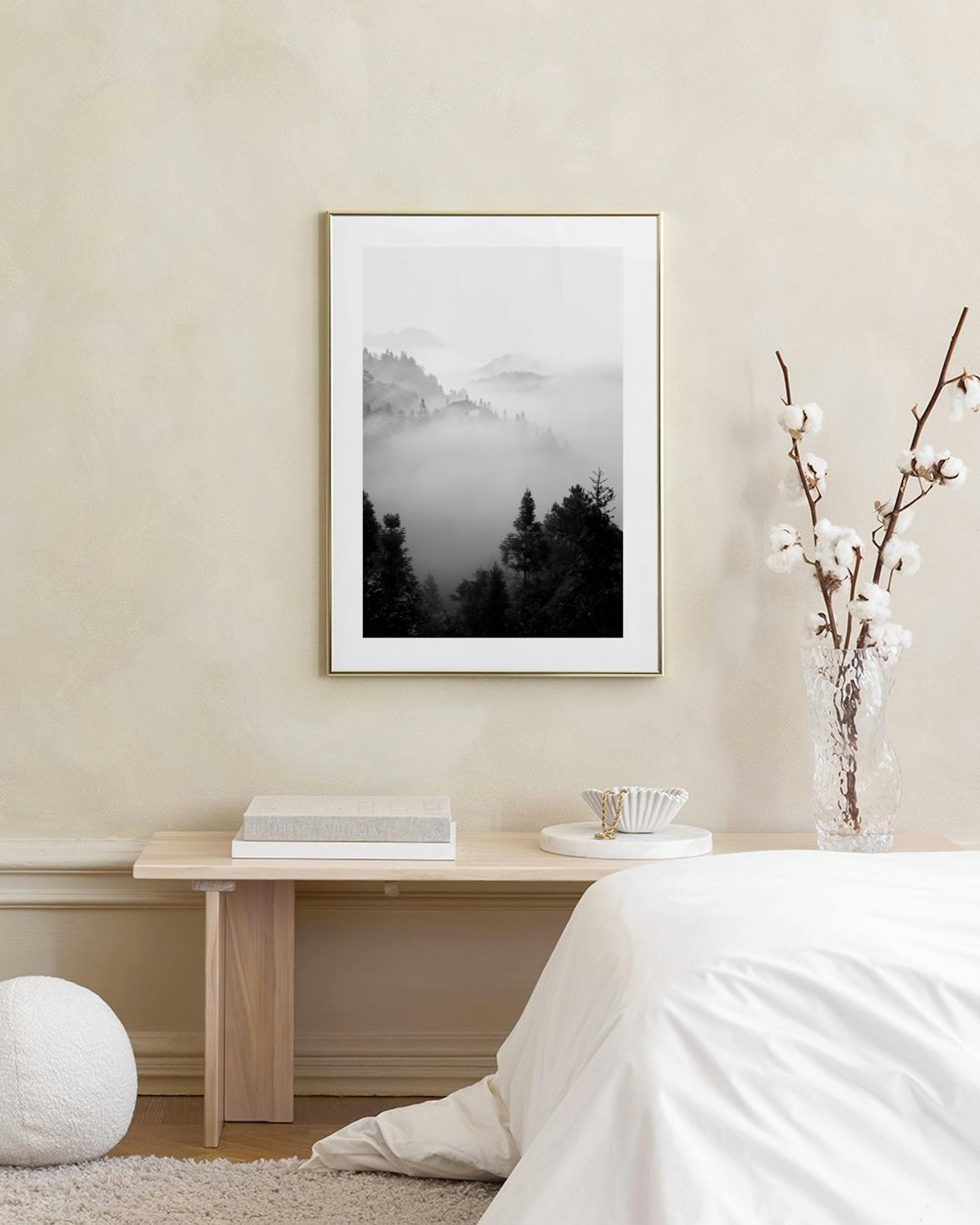Póster monocromático de montañas cubiertas de niebla y árboles oscuros, exhibido en una pared gris junto a una silla y un jarrón