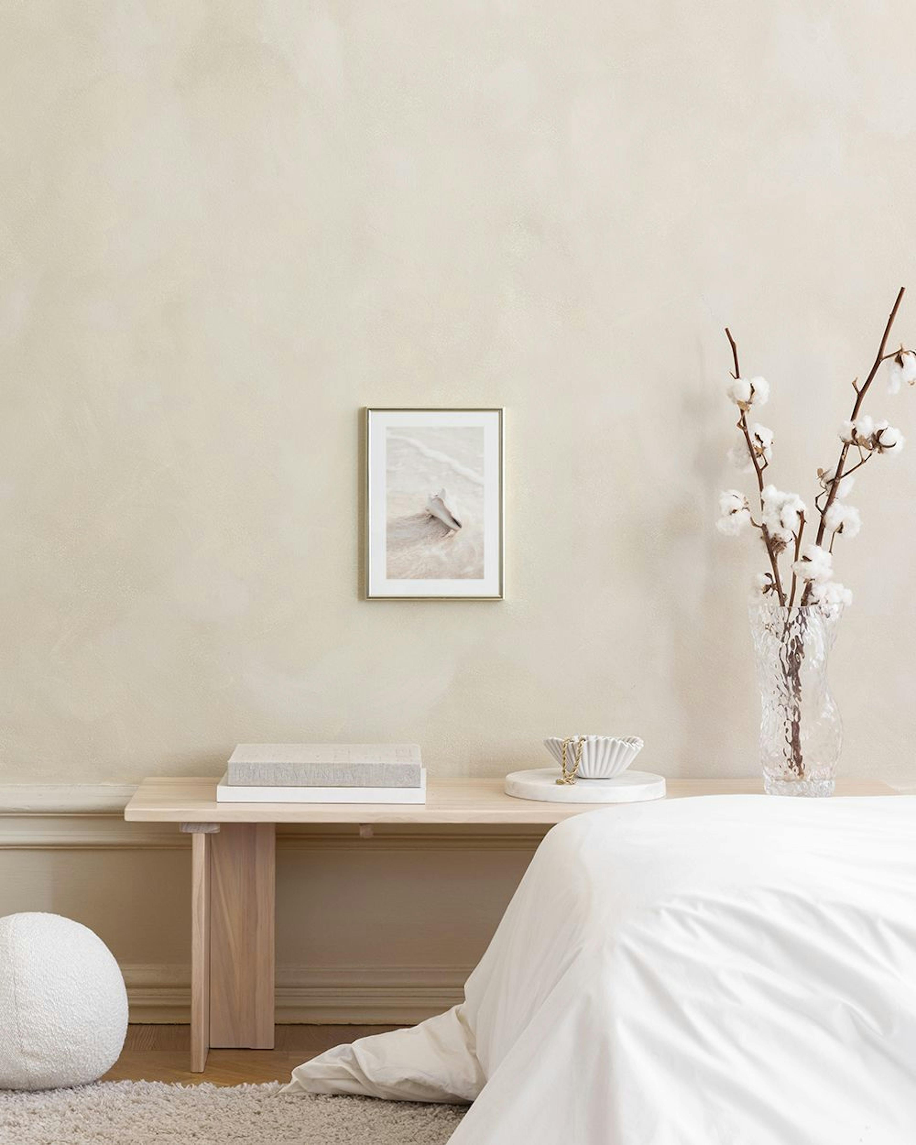 A print of a conch shell on a sandy beach with gentle waves, hanging above a bedside table with a plant and bed.