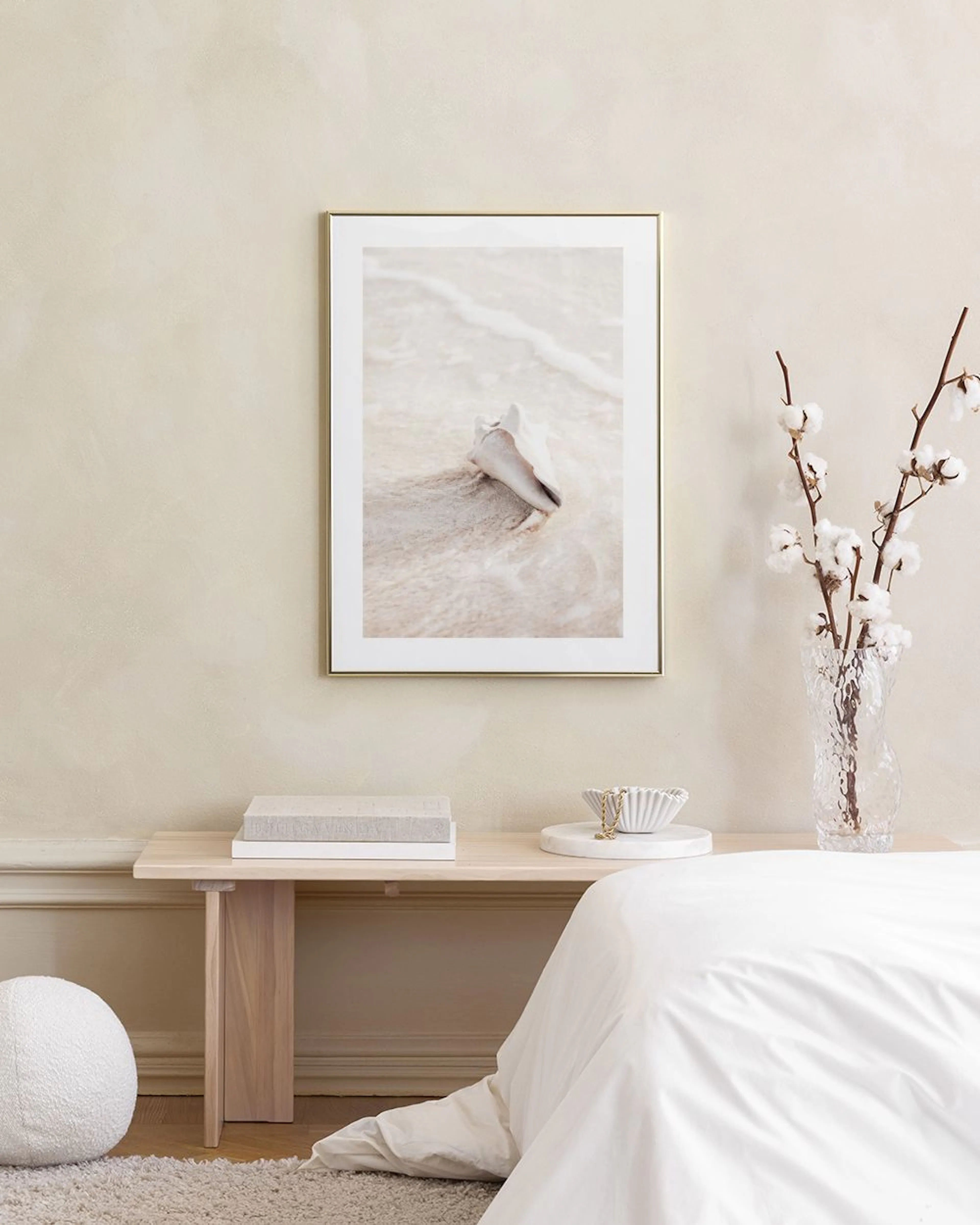 A print of a conch shell on sand with gentle waves, hanging above a concrete side table beside a bed in a bright room.