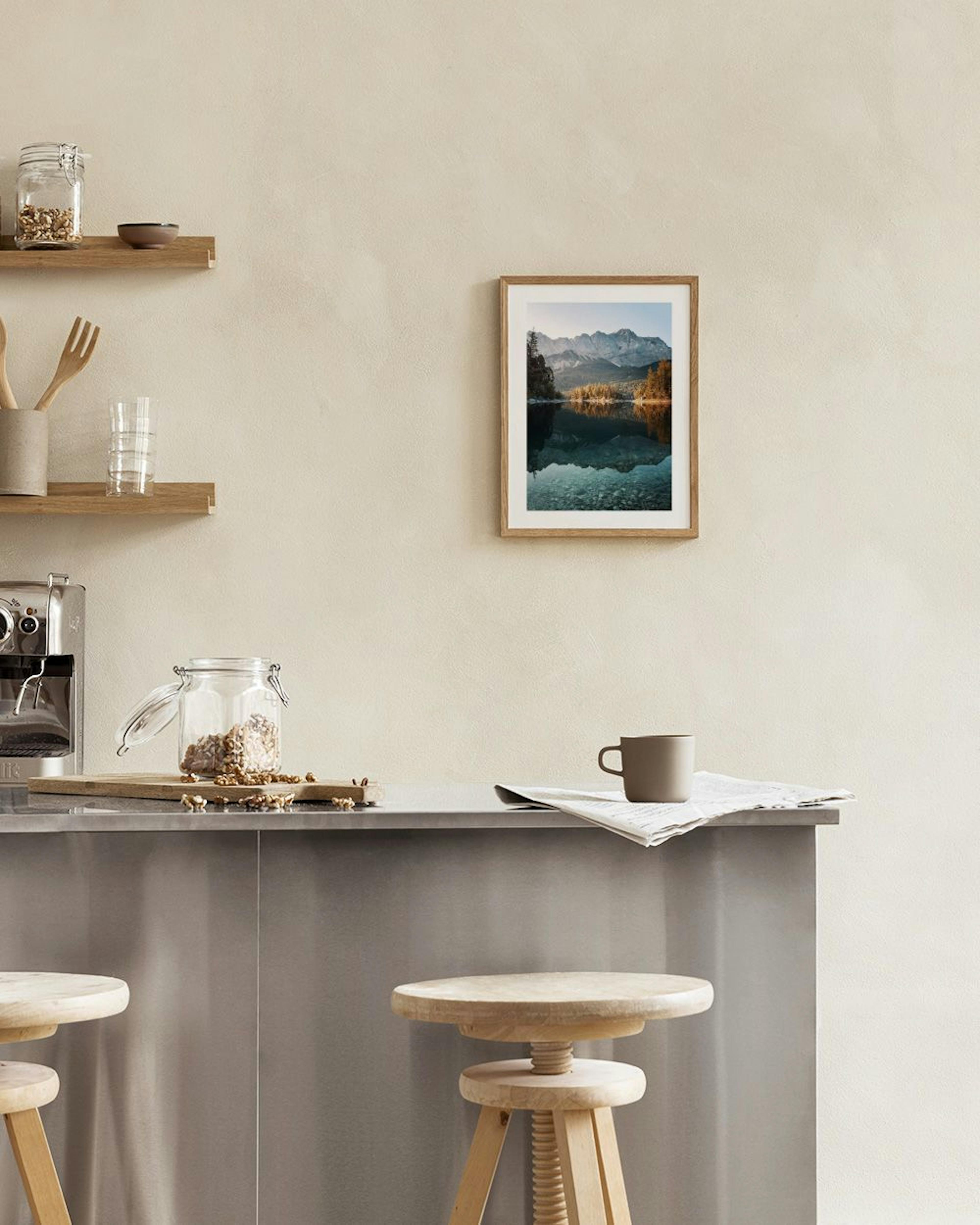 A print of a serene lake reflecting mountains and trees, hanging on a grey wall above a white side table with two vases and book