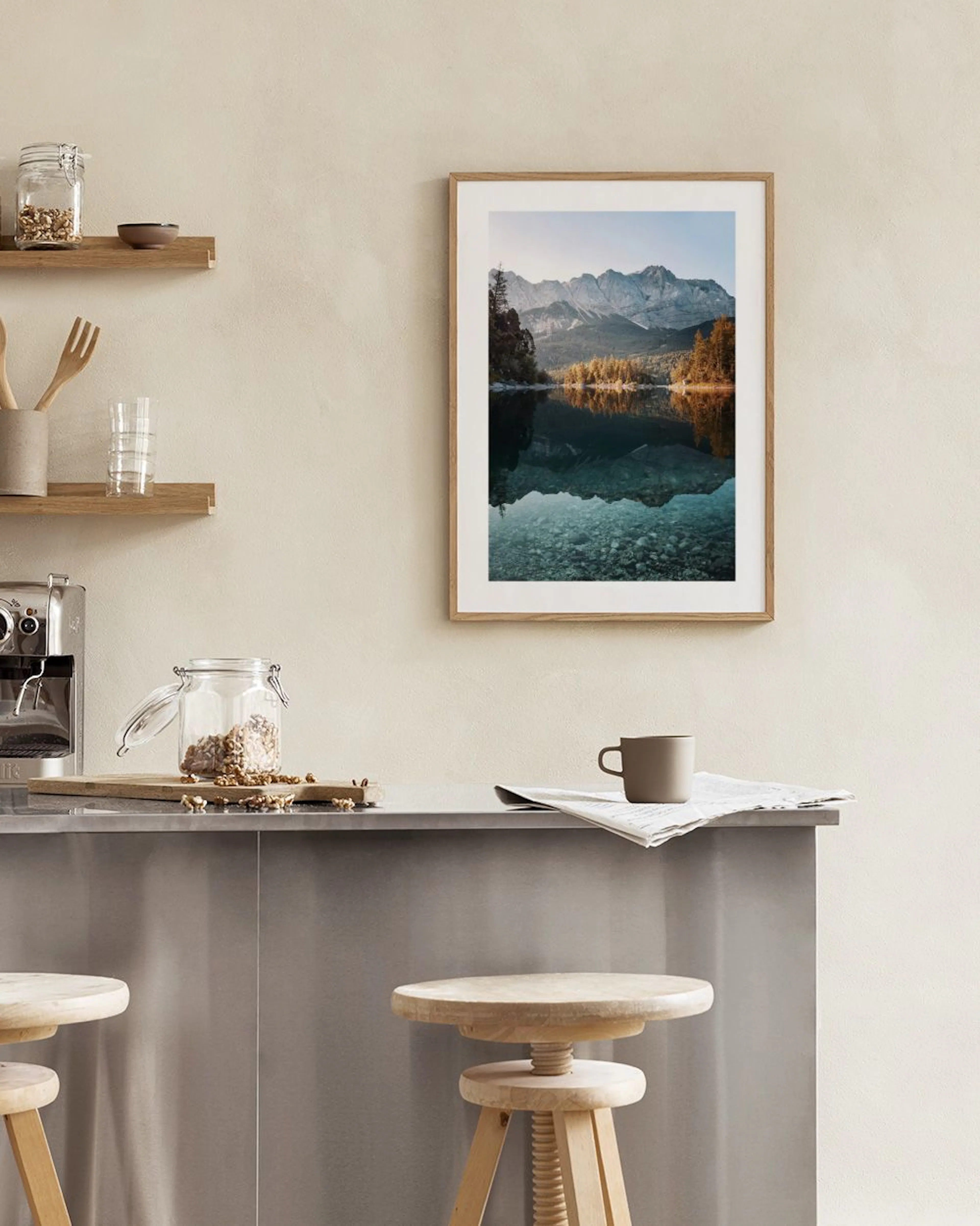 A print of a clear lake reflecting mountains and autumn trees, hanging above white decorative objects on a table.