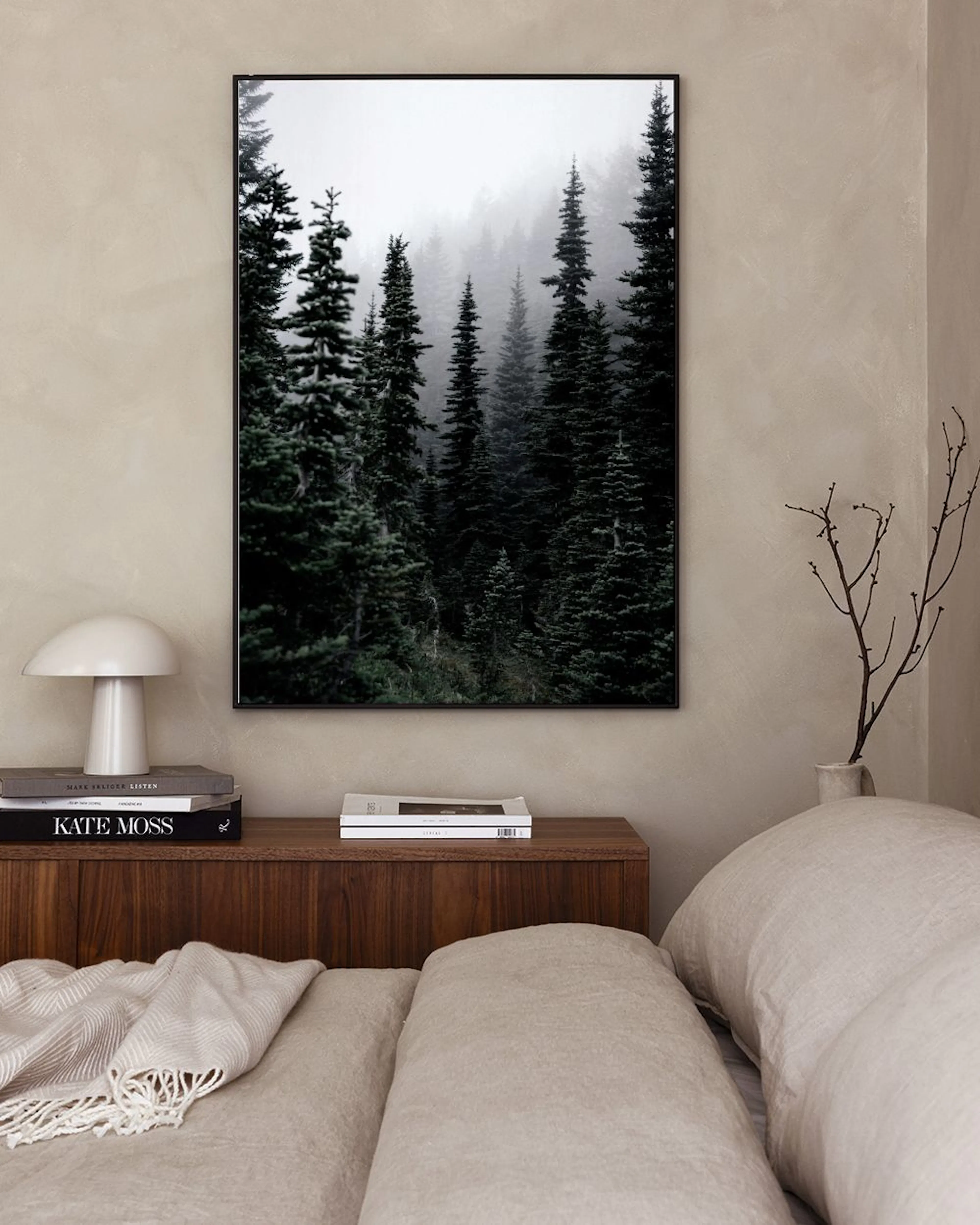 A print of a dark green forest with fog hanging in a white room with a green chair and a white side table.
