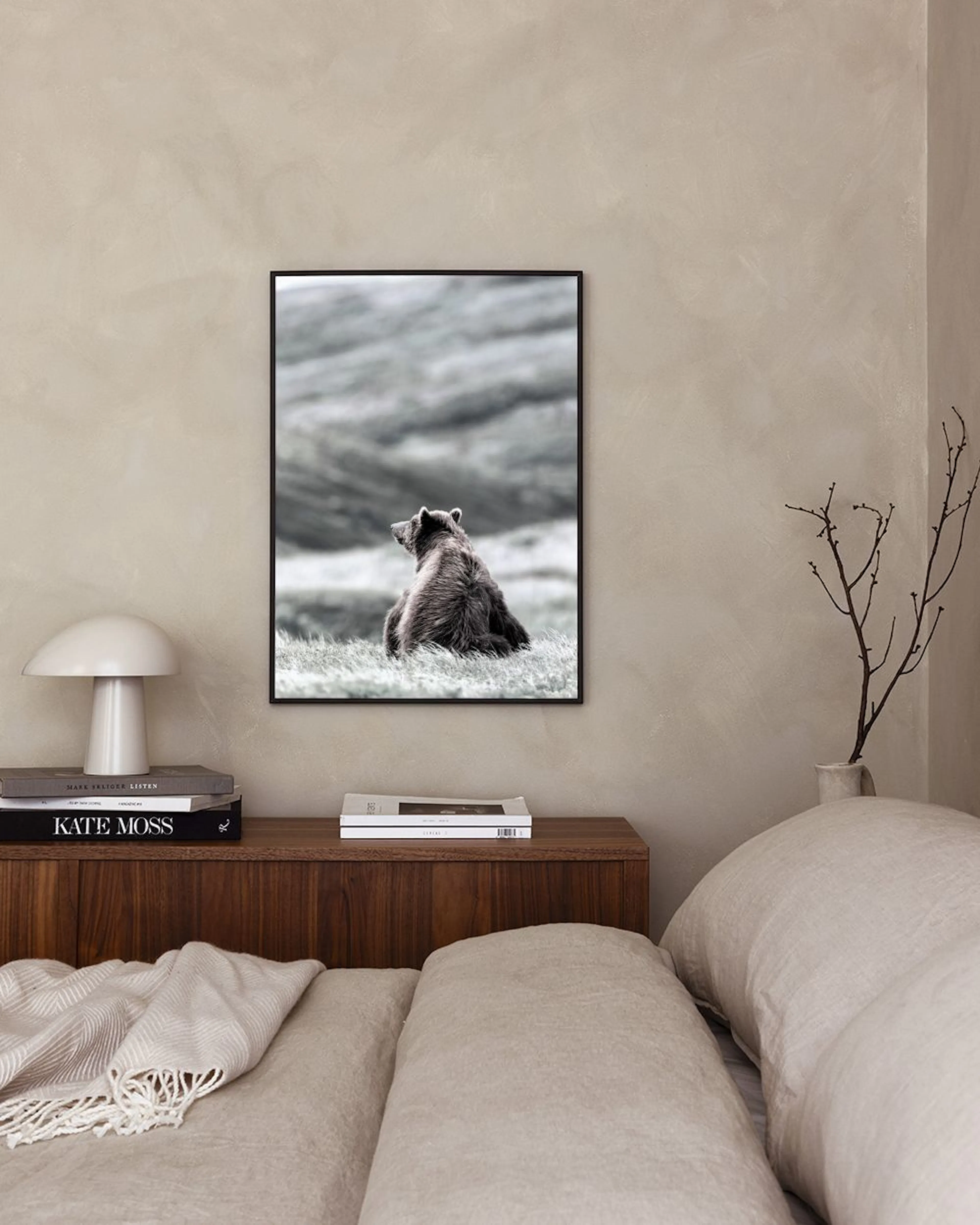 Affiche dun ours brun assis dans un champ dherbe, vue de dos, affichée sur un mur blanc au-dessus dun tabouret avec des livres.