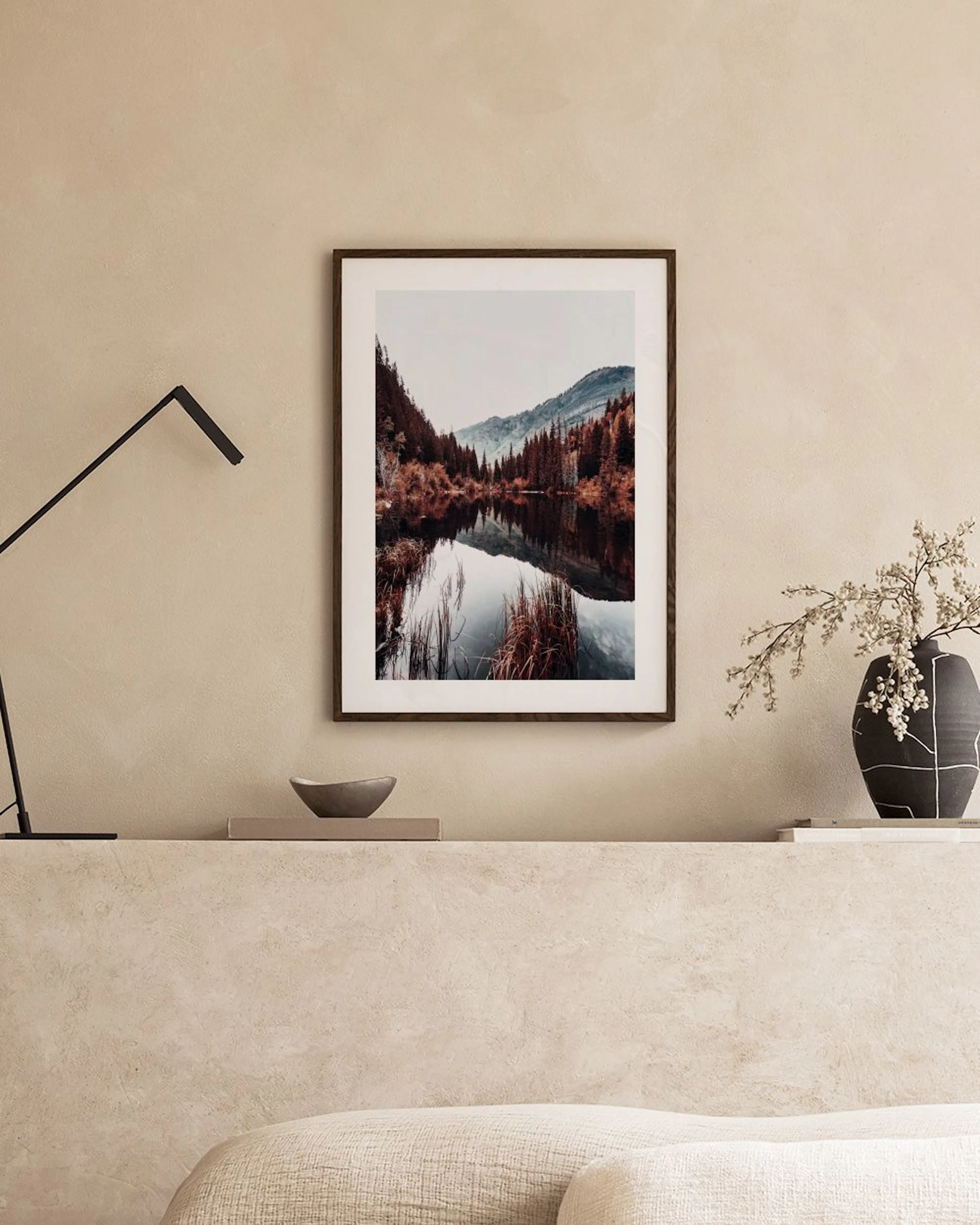 Plakat af en fredfyldt sø omgivet af efterårstræer og bjerge, hængende over et skrivebord med en vase og bøger.