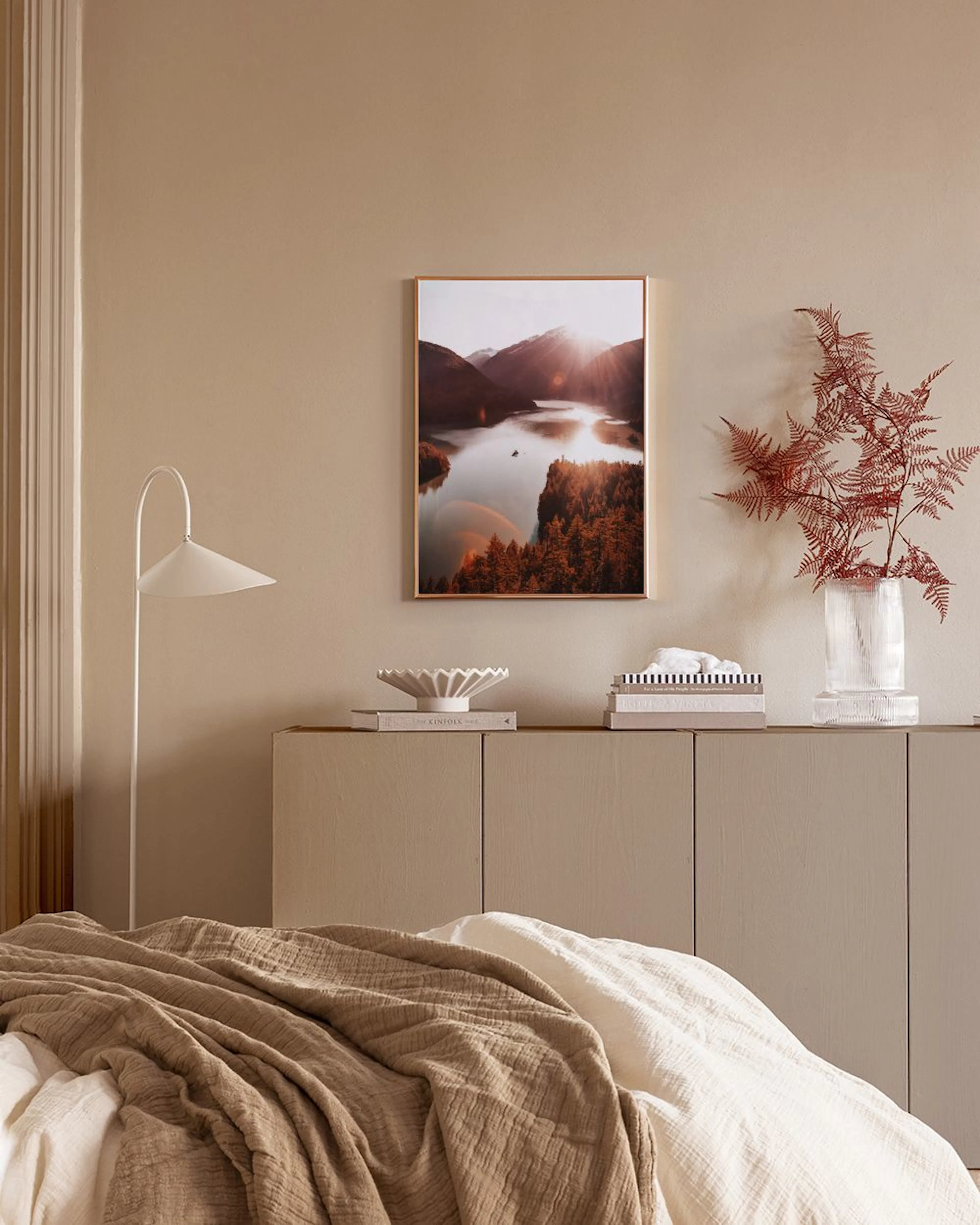 Poster av en sjö omgiven av berg och höstträd, med solstrålar som reflekteras i vattnet, hängande i ett ljust rum.