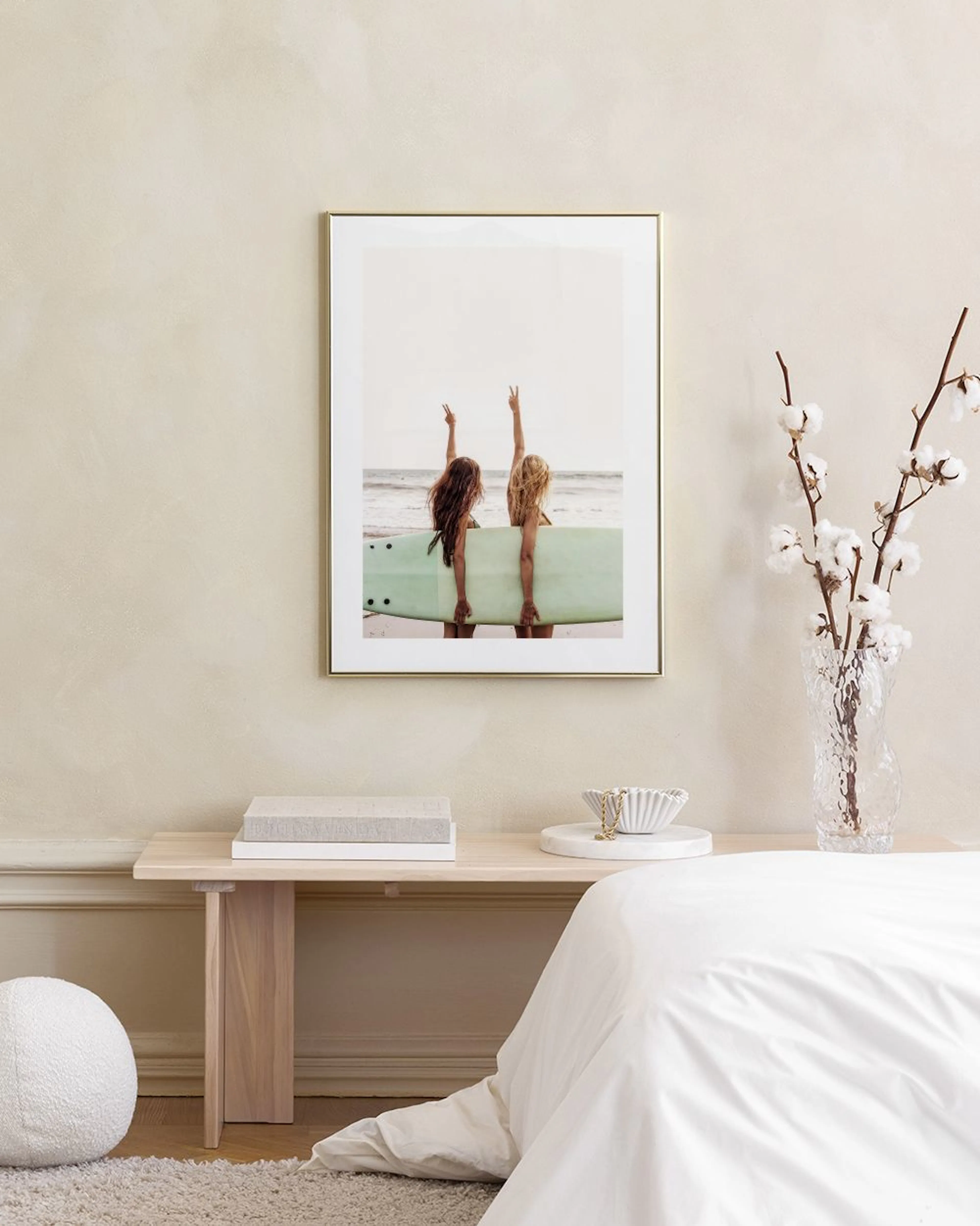 A print of two surfers with a light green surfboard, hands raised in peace signs, hanging above a white console table with books