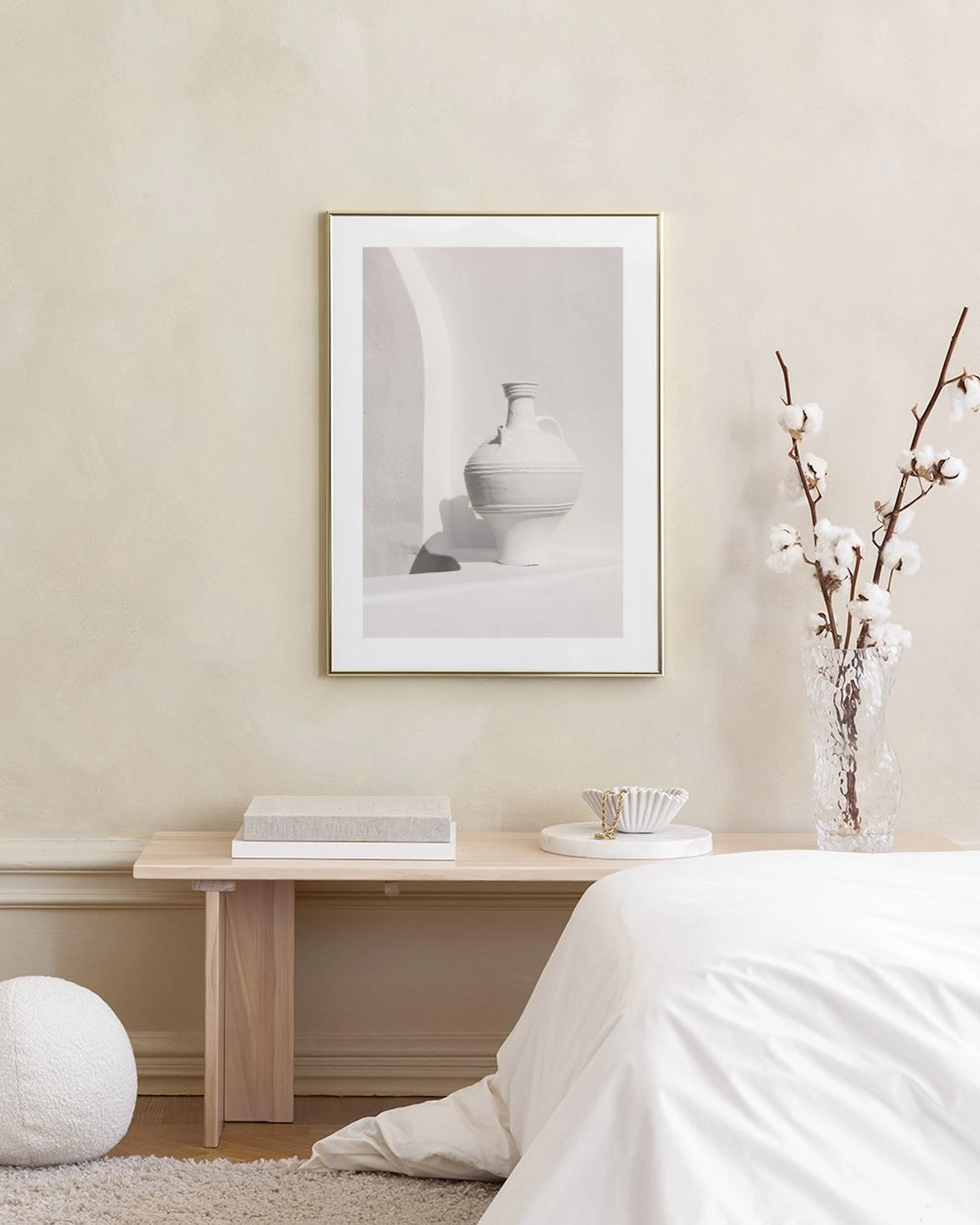 A print of a white rustic vase with handles in black and white, hanging above a wooden bench in a living room.
