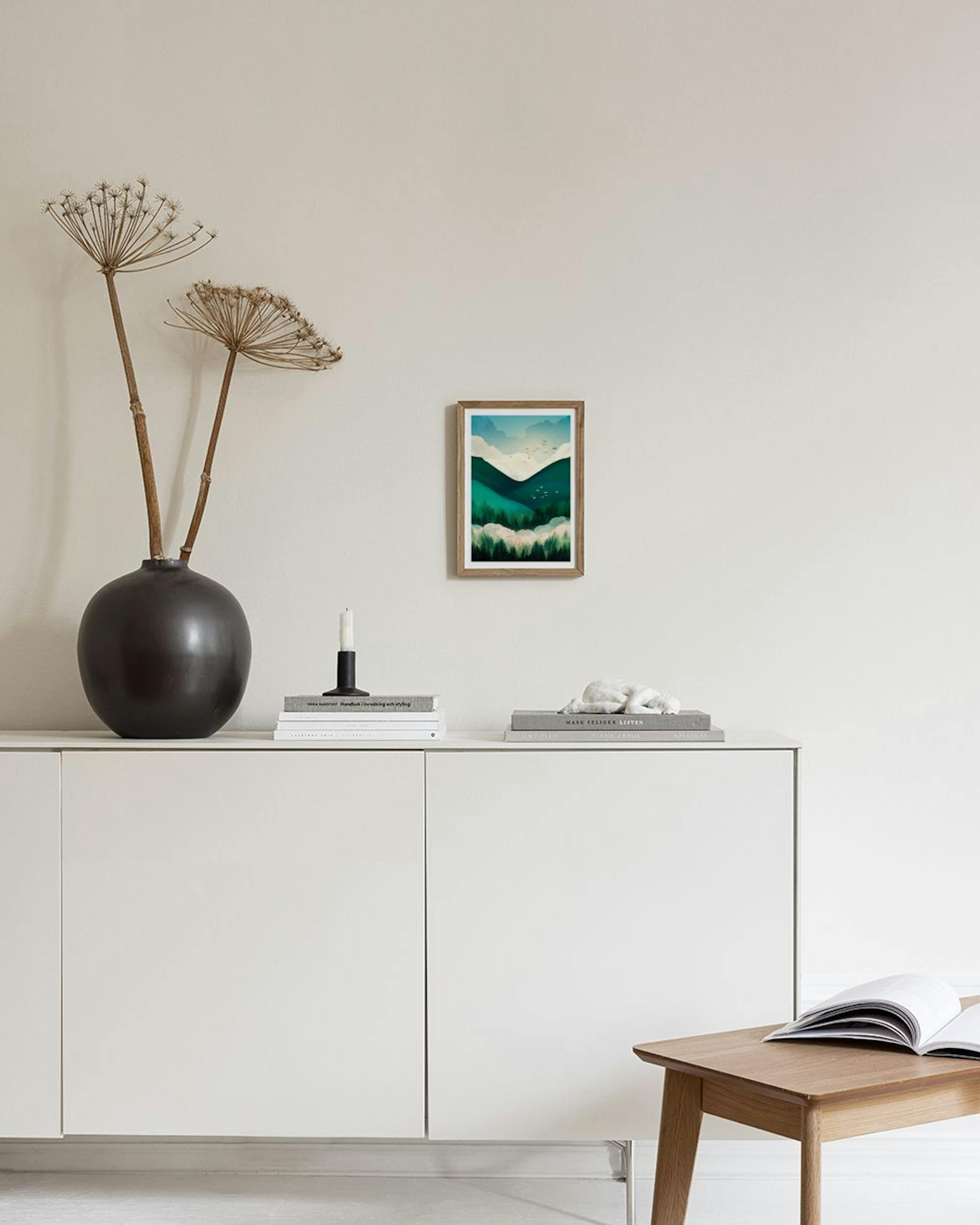 Poster de paisagem montanhosa em tons de verde e azul, com árvores e aves, pendurado acima de um móvel branco com um vaso e livr