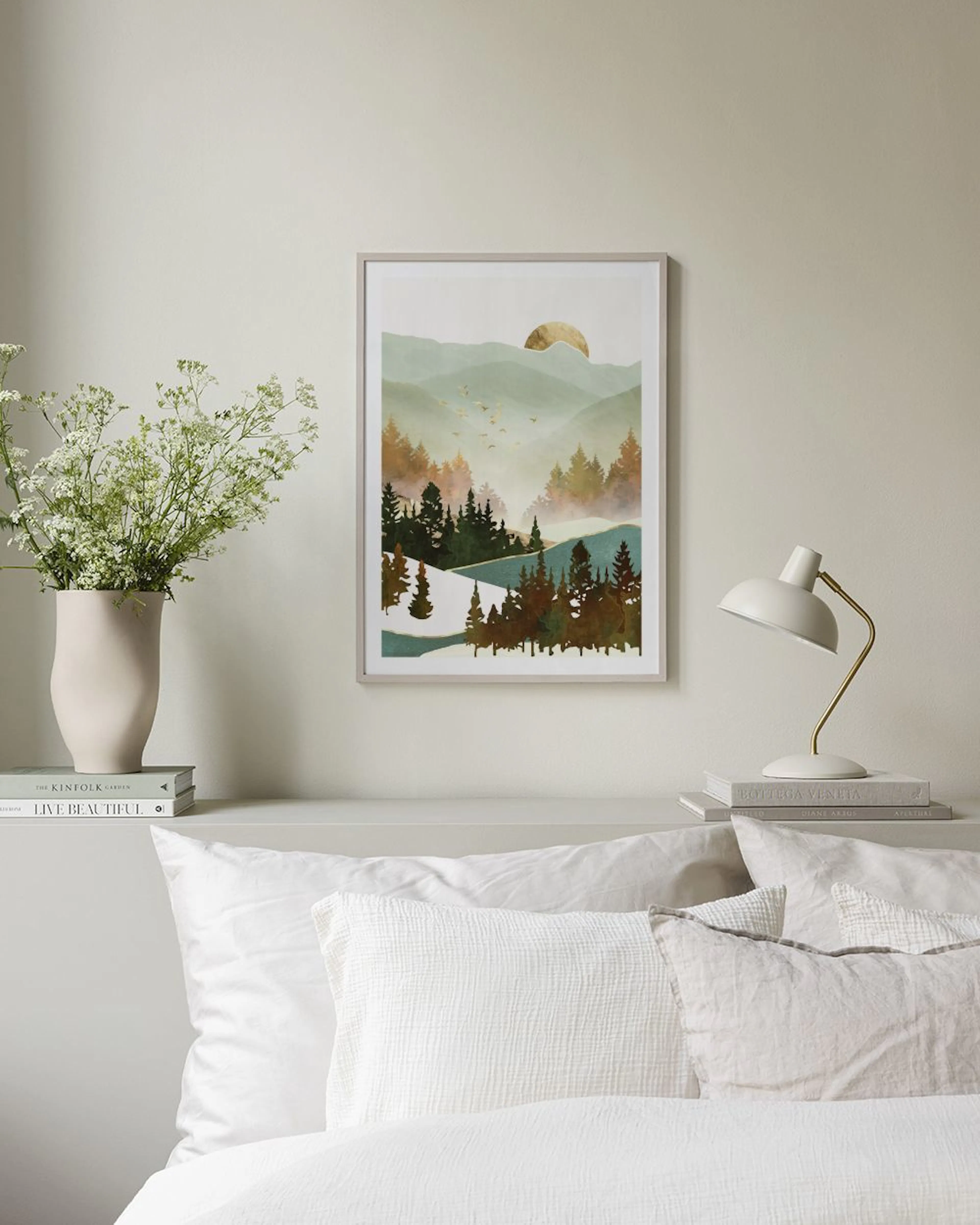 Plakat med et bjerglandskab ved solnedgang med træer og fugle, hængende på en væg over en redt seng.