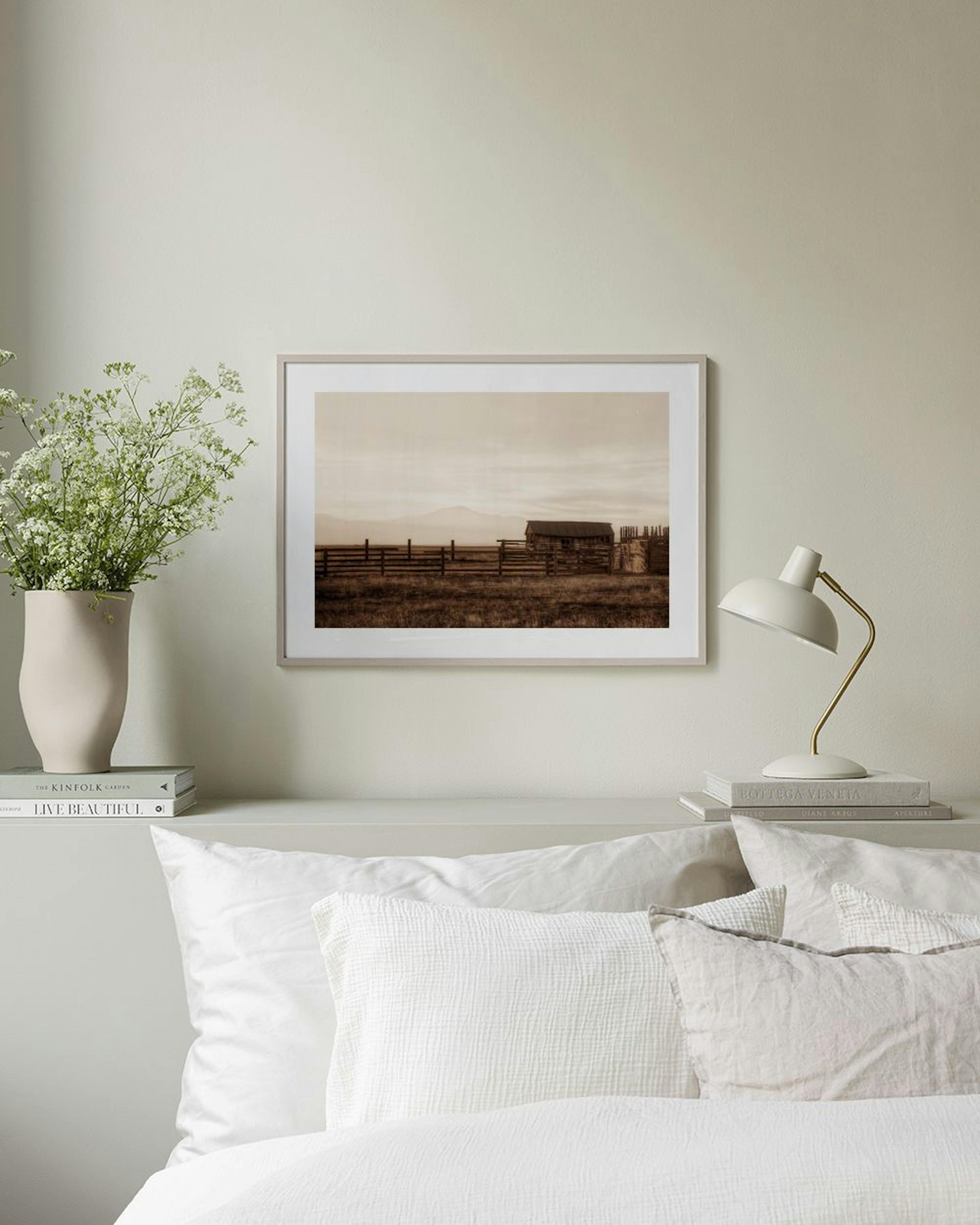 Póster de una cabaña rústica y una cerca bajo un cielo brumoso, exhibido sobre un mueble de madera con jarrones y libros.