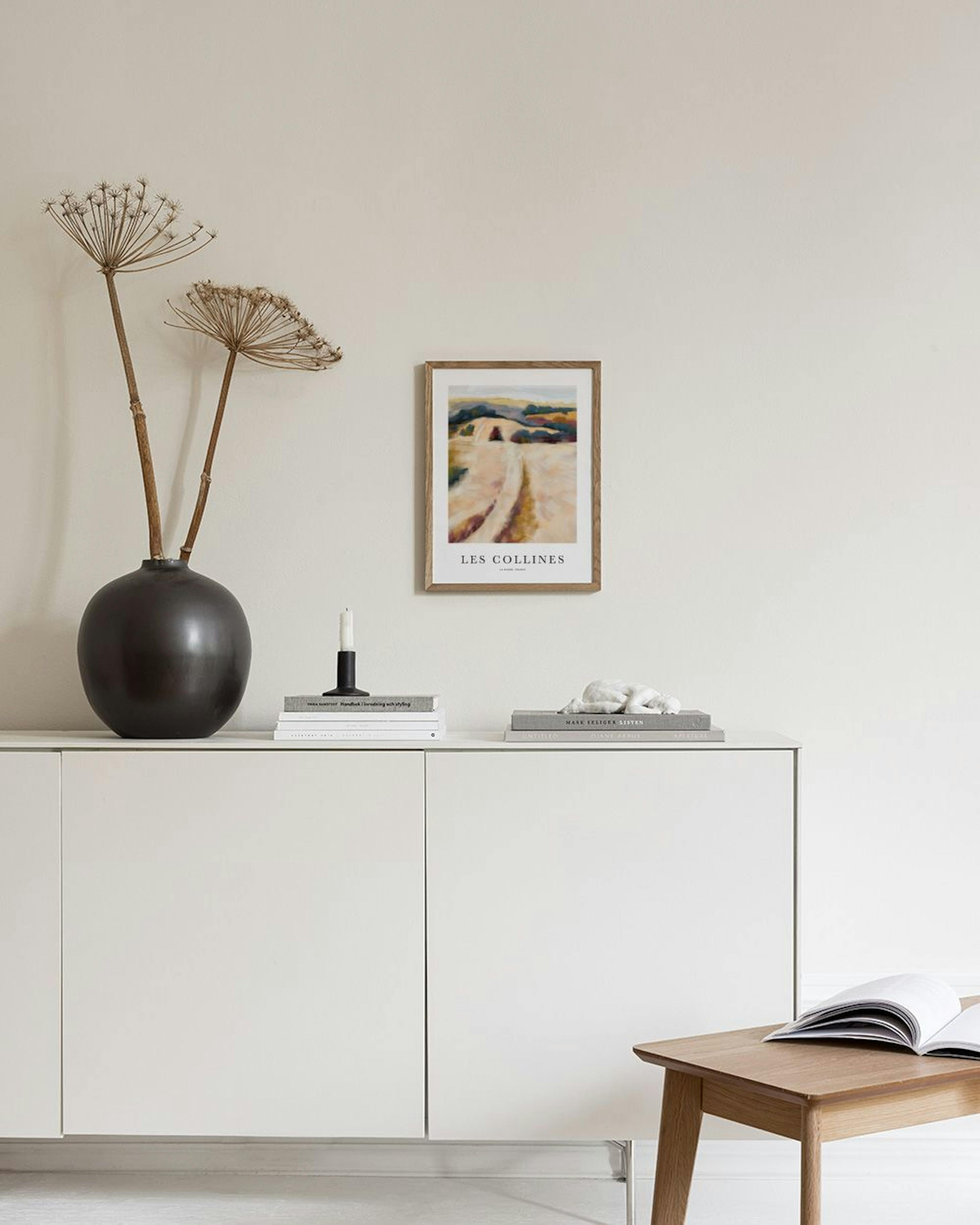 Póster de paisaje con colinas en tonos tierra, azul y ocre, colgado sobre un sillón beige en una sala moderna.