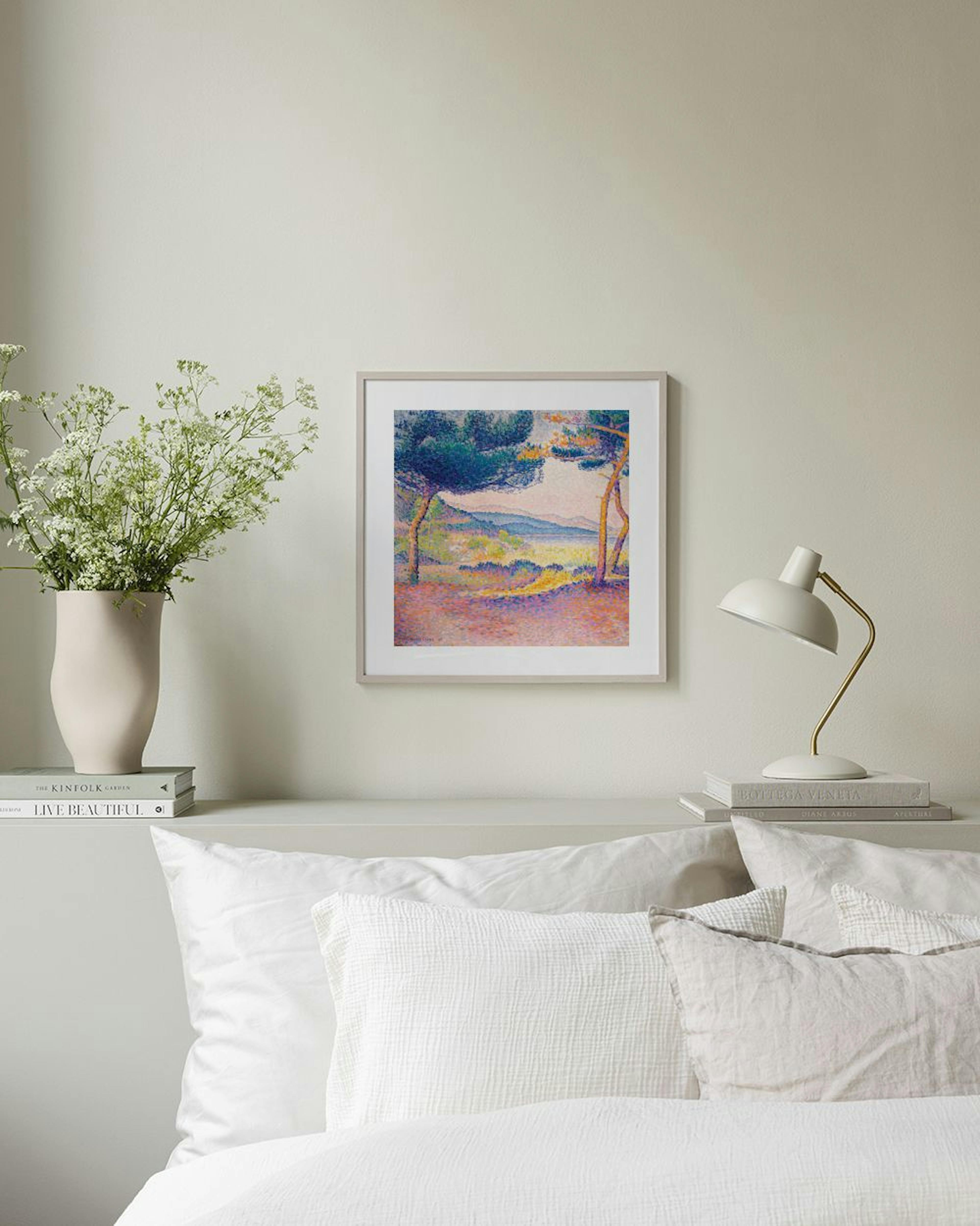 A pointillist landscape print with green trees and a coastal view, displayed above a light wood desk with books and decorative o