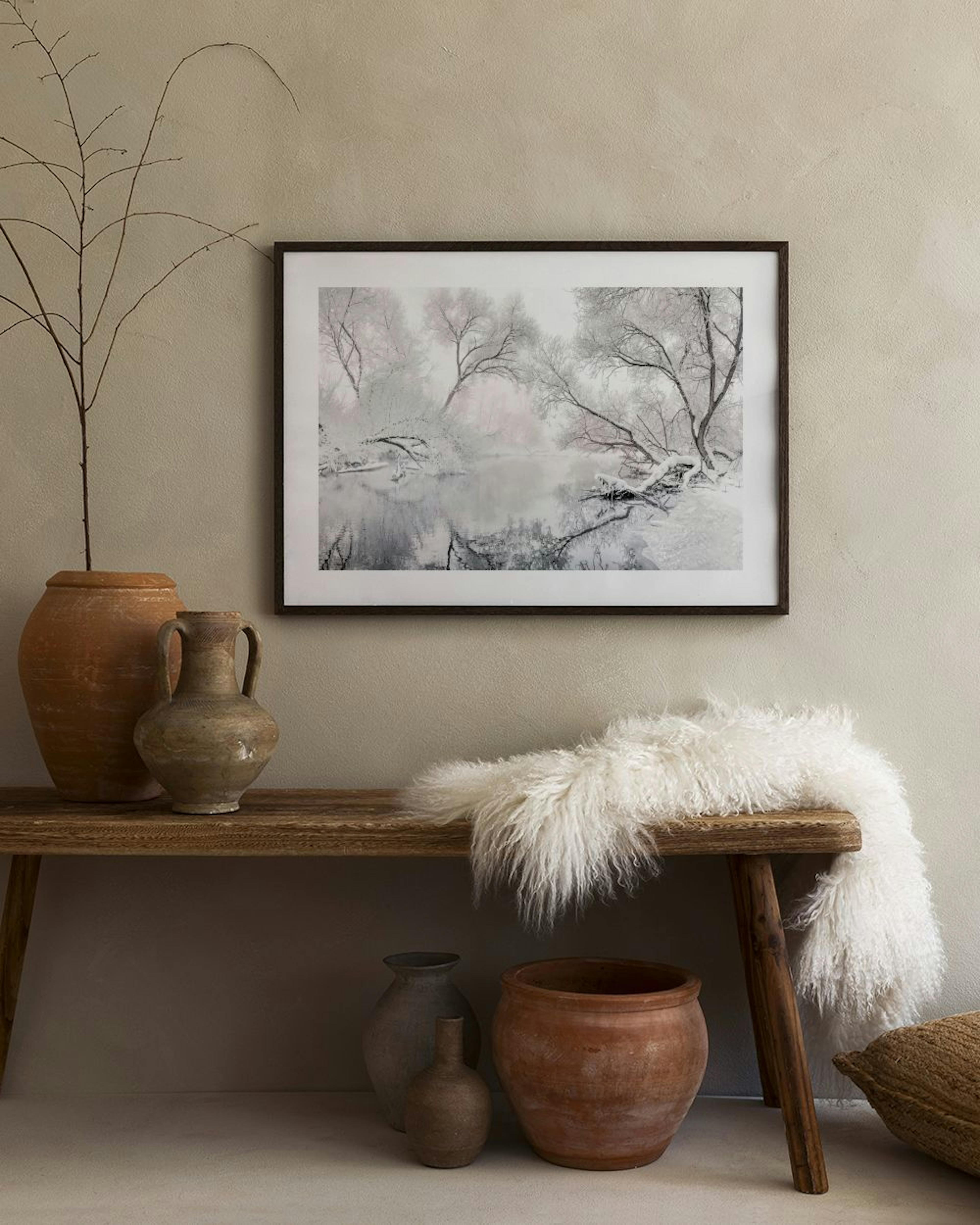 A winter landscape print with snow-covered trees and a misty lake, hanging above a wooden cabinet with decorative vases and book