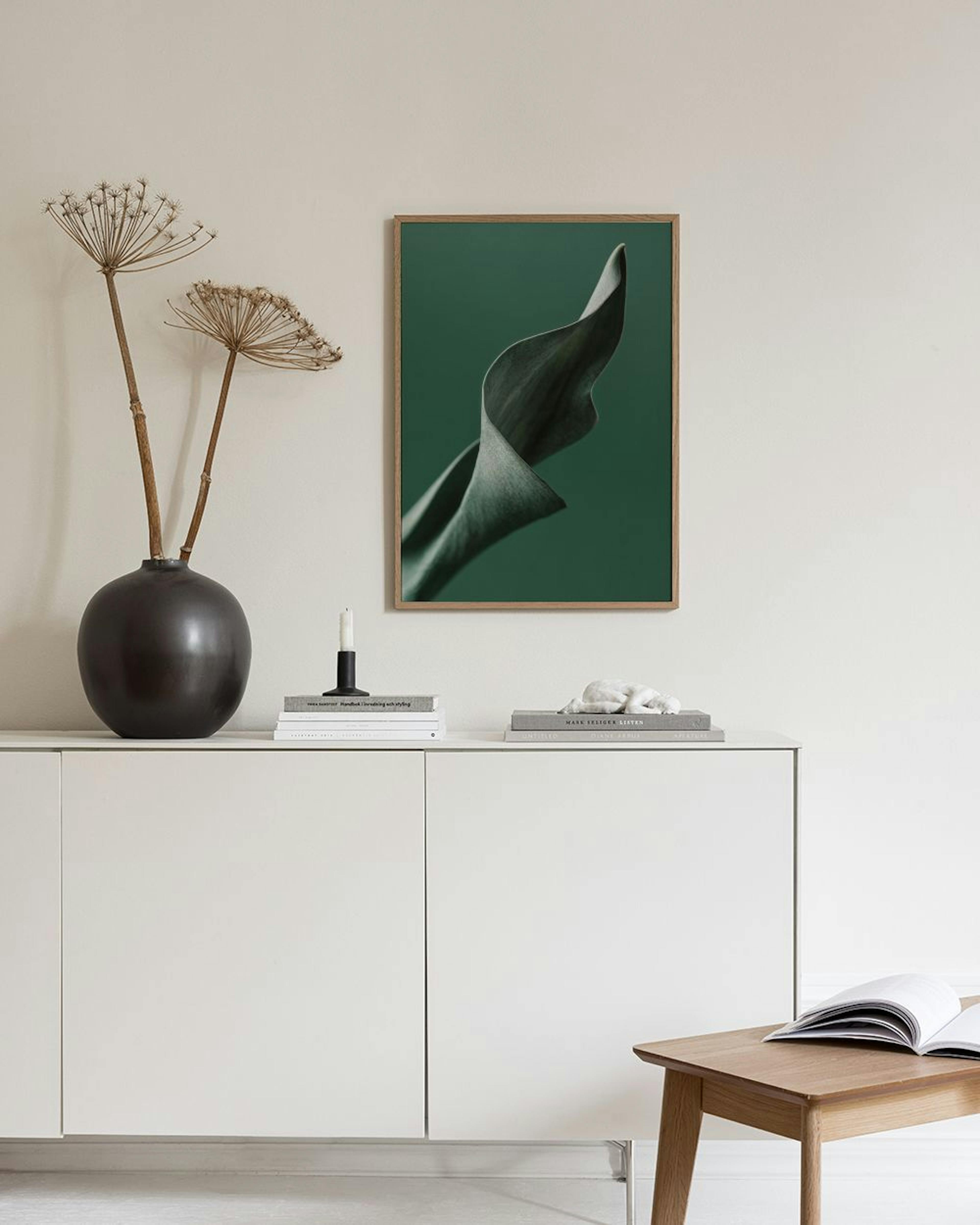 Póster de una hoja verde oscuro con formas orgánicas y curvadas, colgado en una pared blanca sobre un puf marrón y una mesa auxi