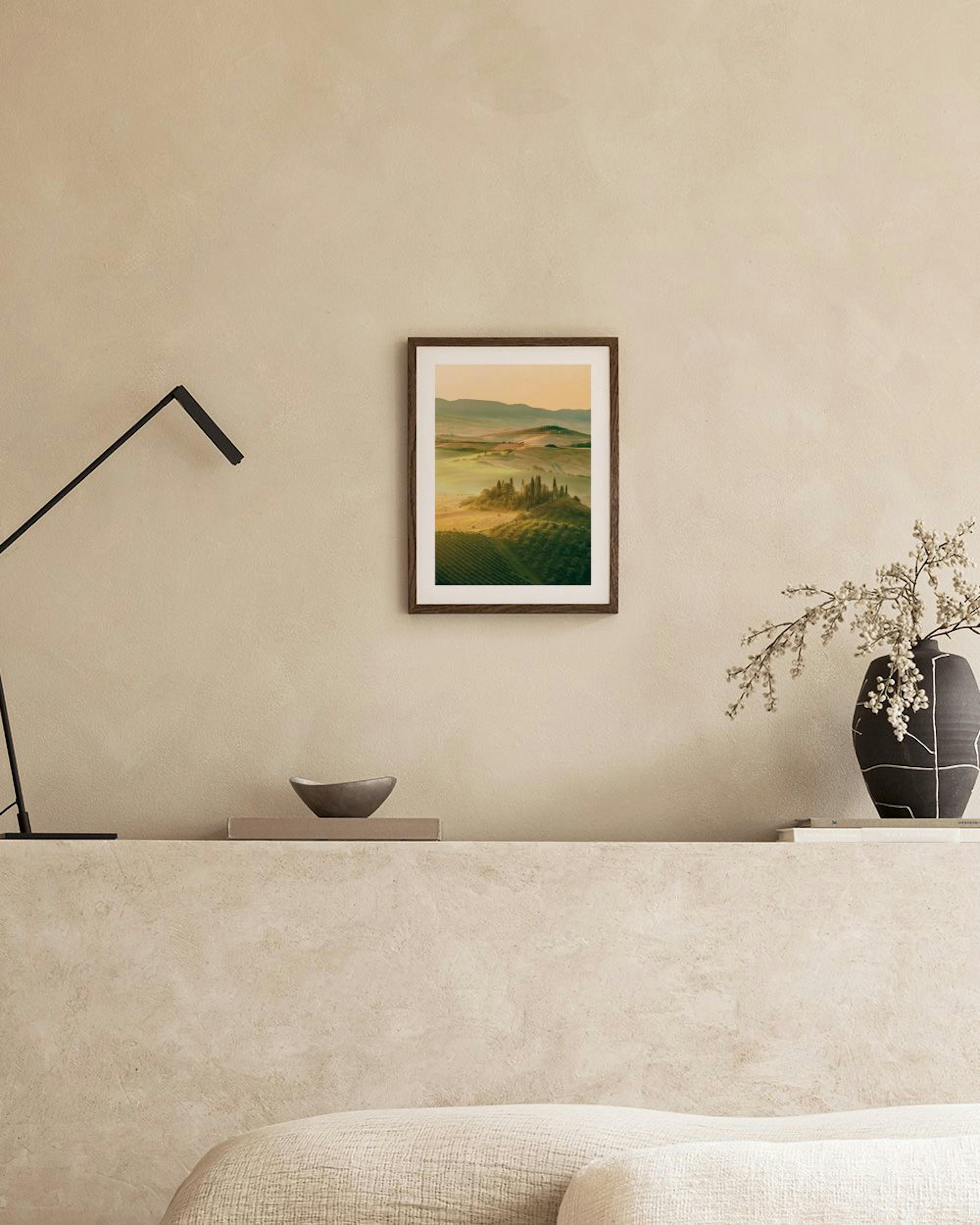 Póster de paisaje toscano con colinas verdes y amarillas, viñedos, y cipreses, exhibido sobre un aparador de mimbre con decoraci
