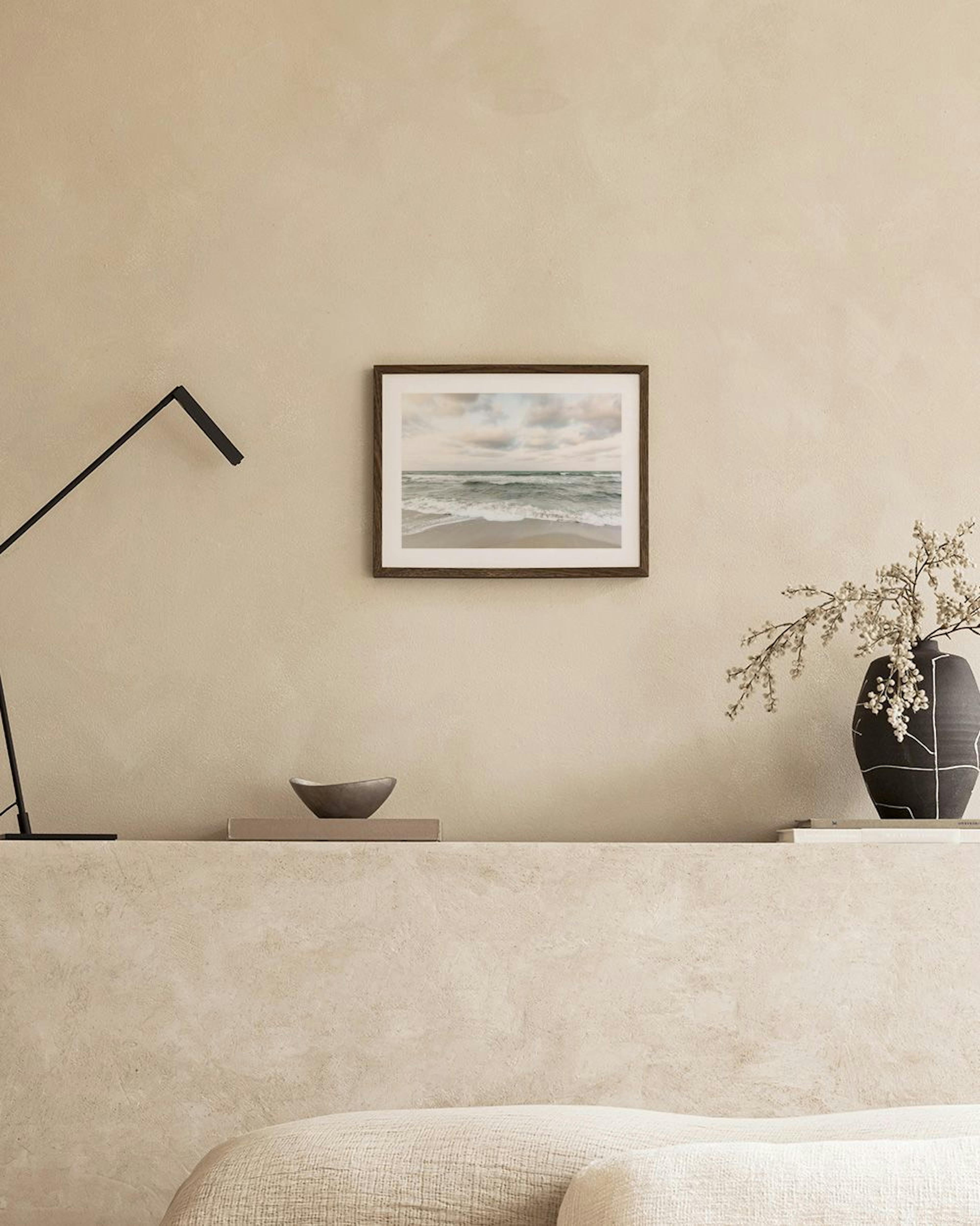 A print of a cloudy ocean beach with waves, hanging above a white console table with a clear vase and decorative objects.