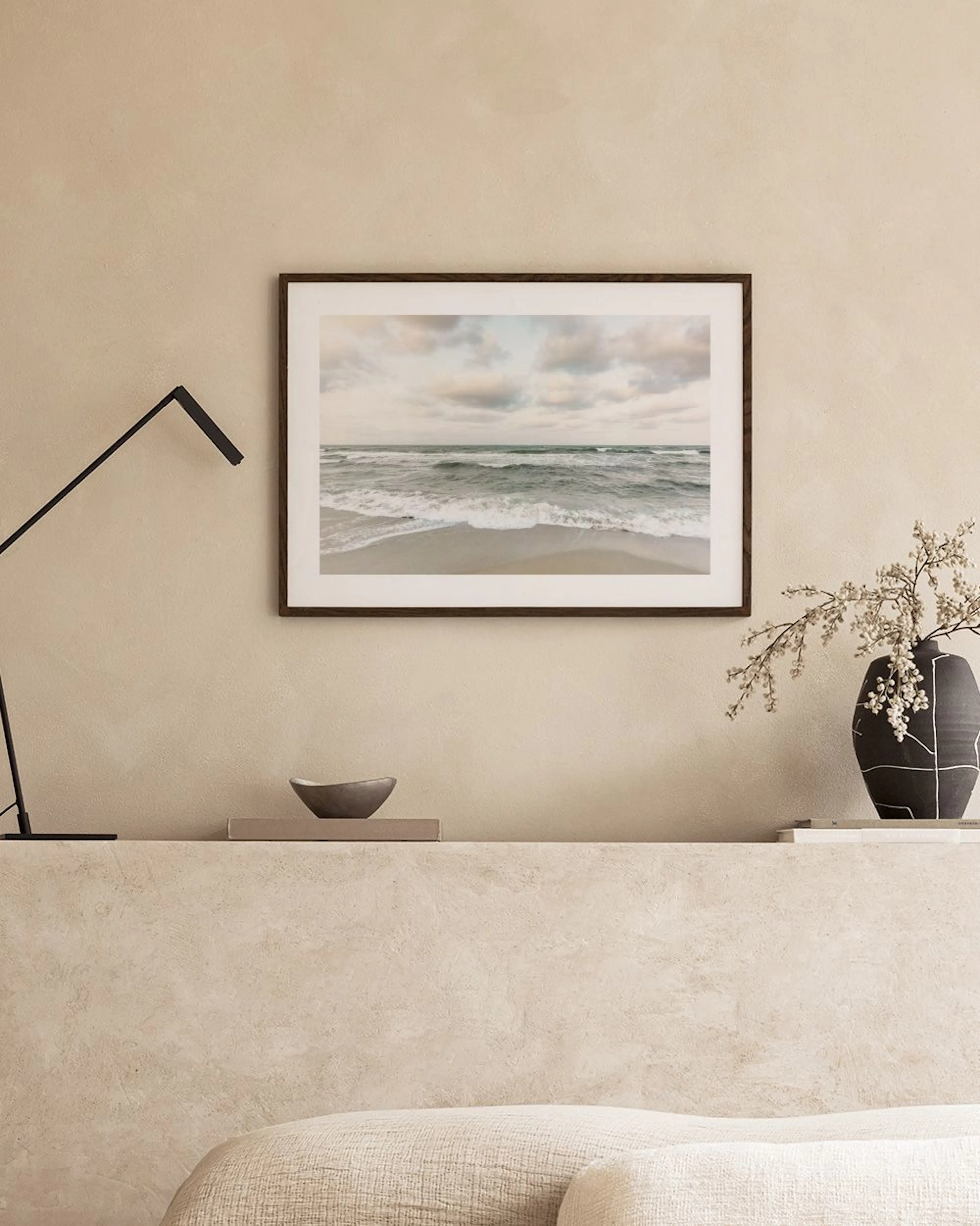 Póster de una playa con olas y cielo nublado, colgado sobre un mueble blanco con una planta y libros.