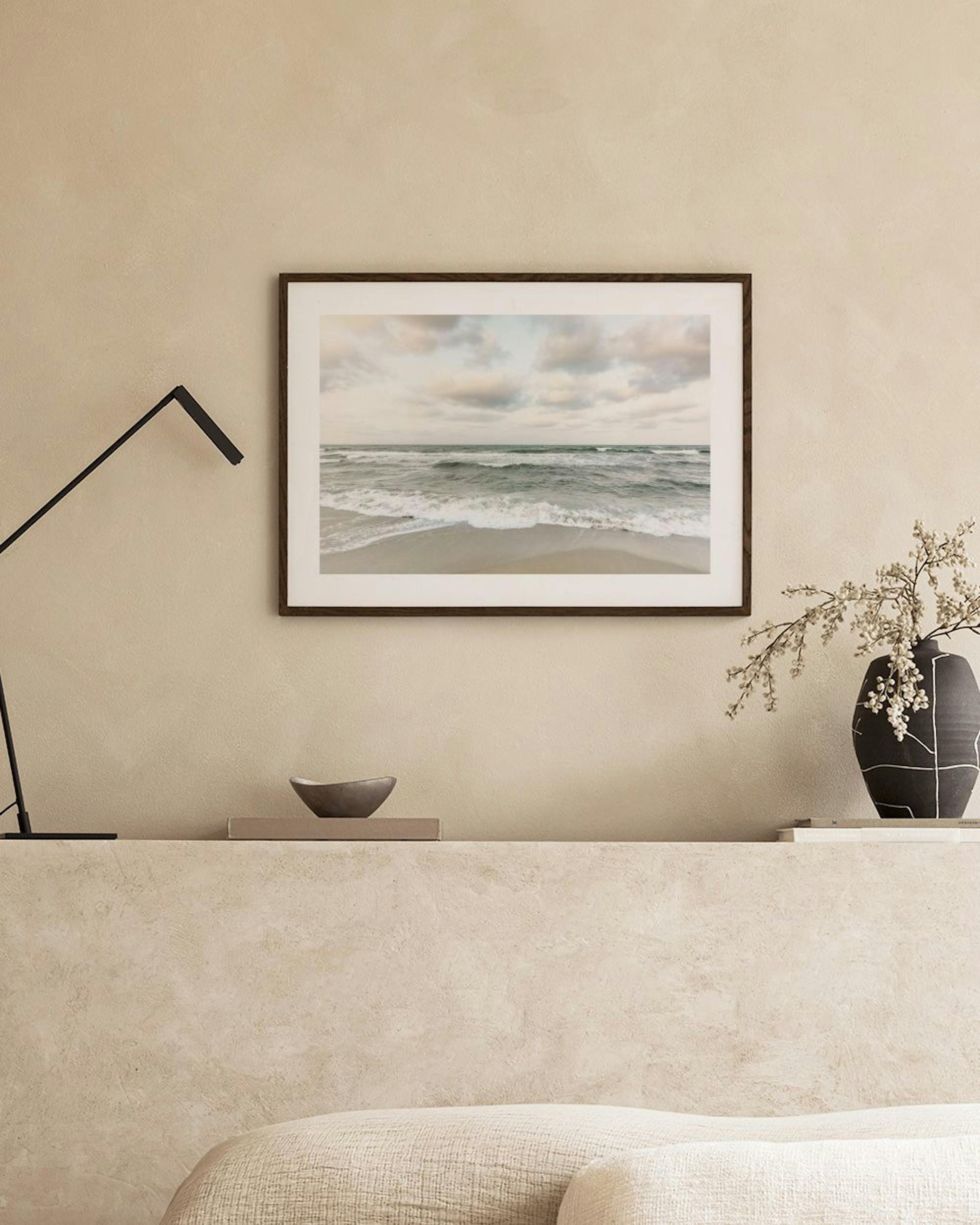 Póster de una playa con olas y cielo nublado, colgado sobre un mueble blanco con una planta y libros.