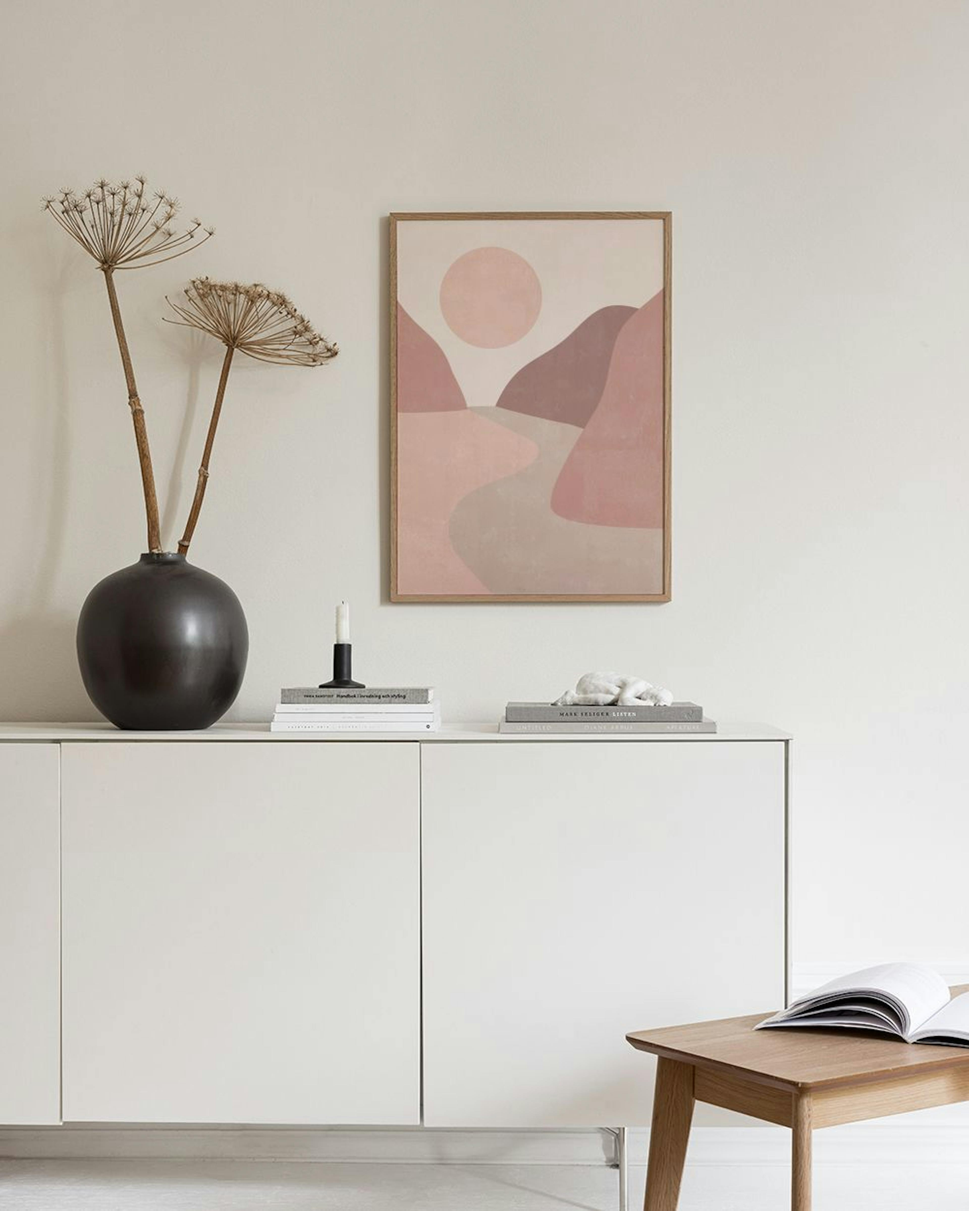Póster abstracto de paisaje con montañas y río en tonos pastel, colgado en una pared clara junto a una silla de madera y un libr