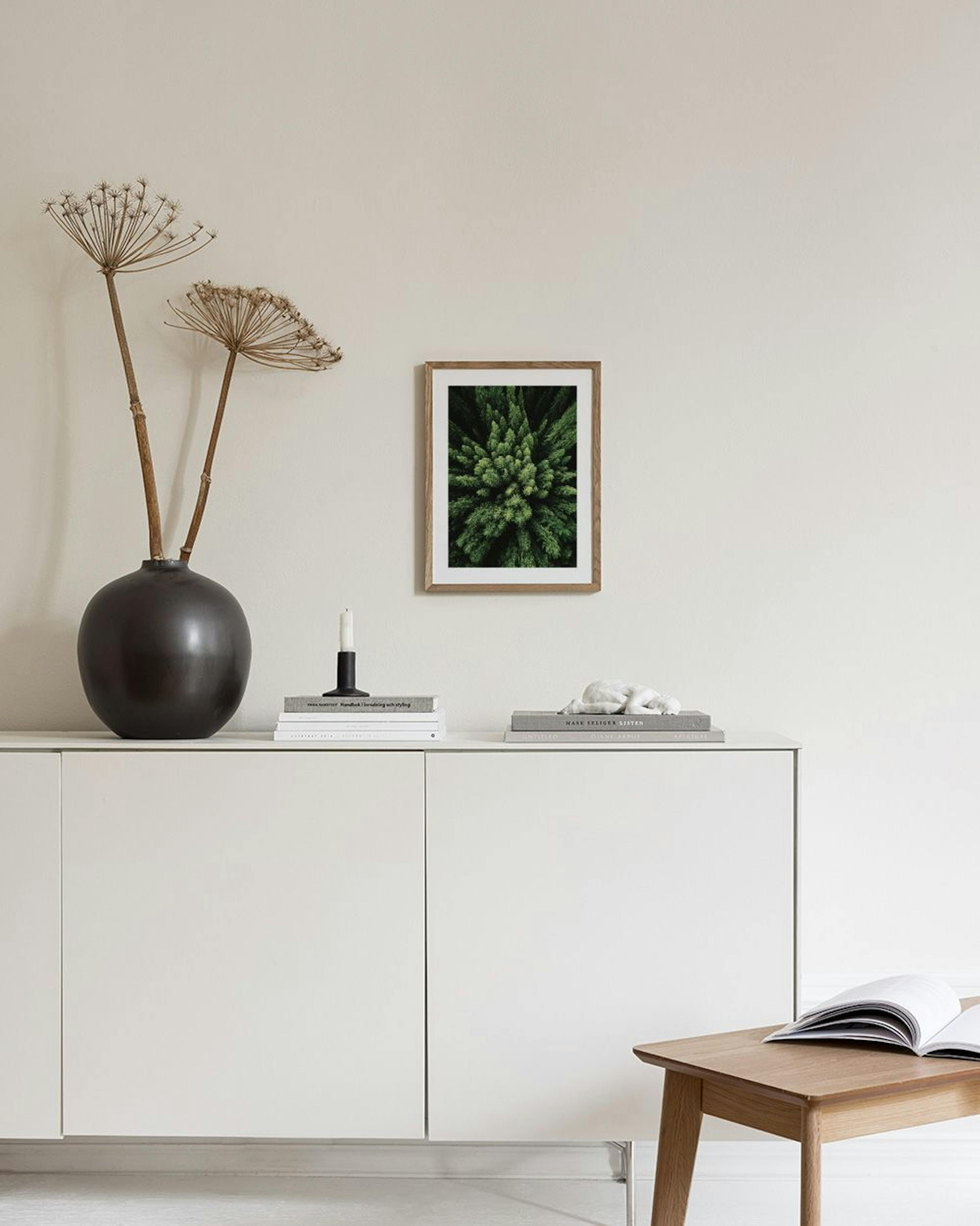 Poster met bovenaanzicht van groene dennenbomen, hangend boven een krukje met boeken in een lichte hal.
